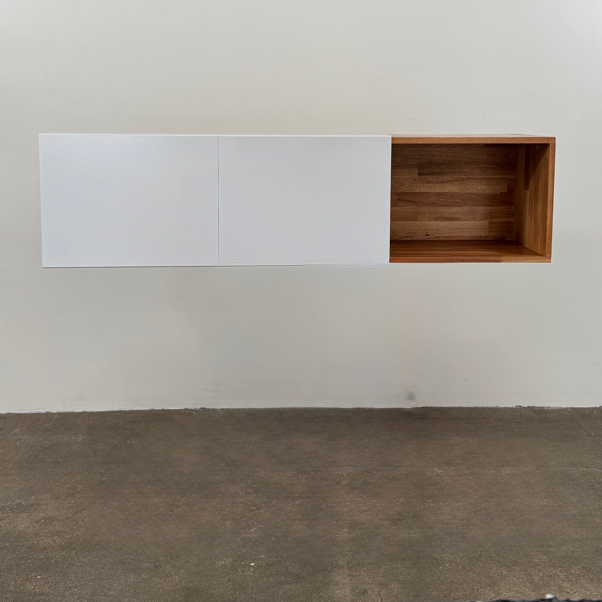 A Modern Resale Mash Studios LAX series 3X Wall Mounted Shelf in natural ash wood and matte white is mounted against a plain wall above a concrete floor, featuring a white exterior with wood-toned interior shelving.