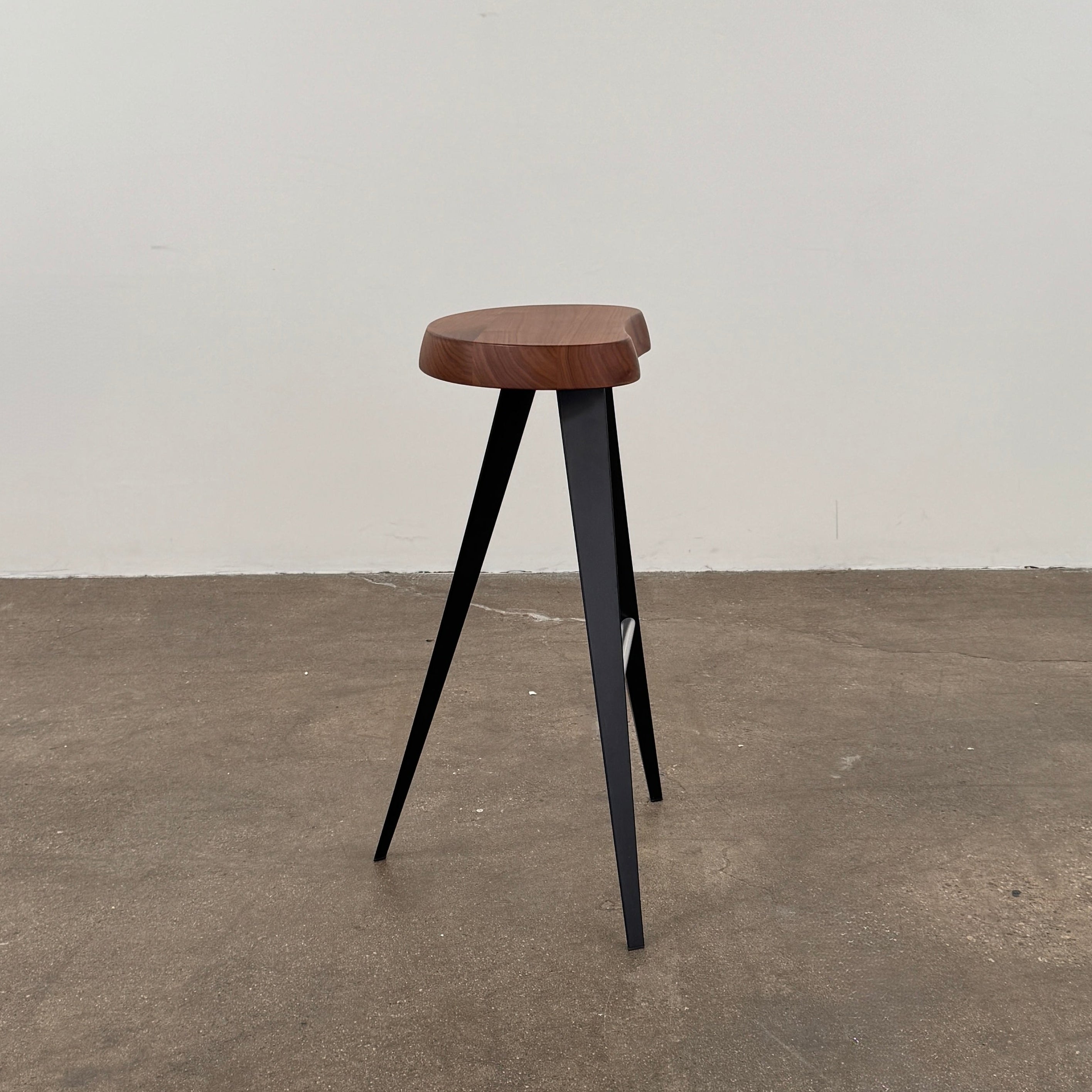 A Cassina Mexique Stool by Cassina, featuring a wooden round seat, three black angled legs, and a metal footrest, is displayed against a plain white background—highlighting its mid-century craftsmanship inspired by Charlotte Perriand.