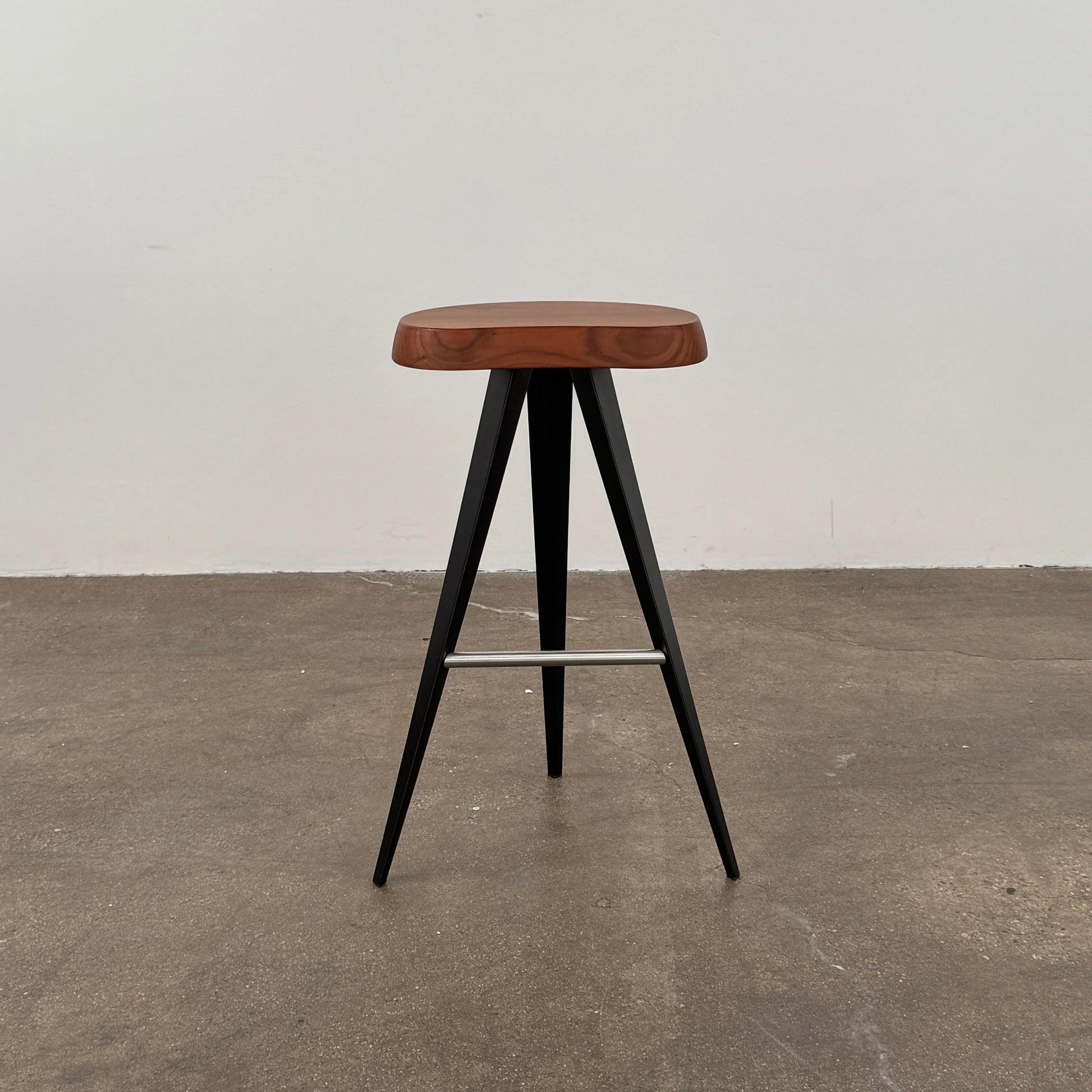 A Cassina Mexique Stool by Cassina, featuring a wooden round seat, three black angled legs, and a metal footrest, is displayed against a plain white background—highlighting its mid-century craftsmanship inspired by Charlotte Perriand.
