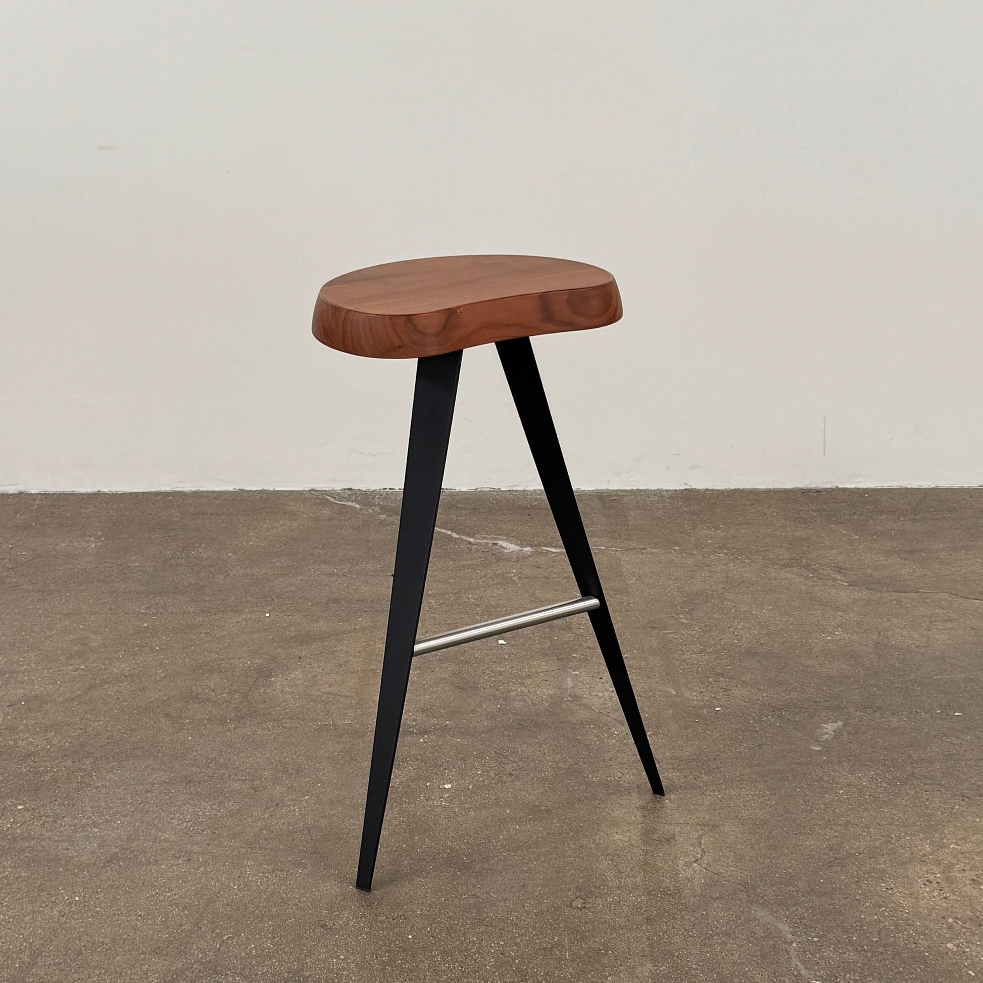 A Cassina Mexique Stool by Cassina, featuring a wooden round seat, three black angled legs, and a metal footrest, is displayed against a plain white background—highlighting its mid-century craftsmanship inspired by Charlotte Perriand.