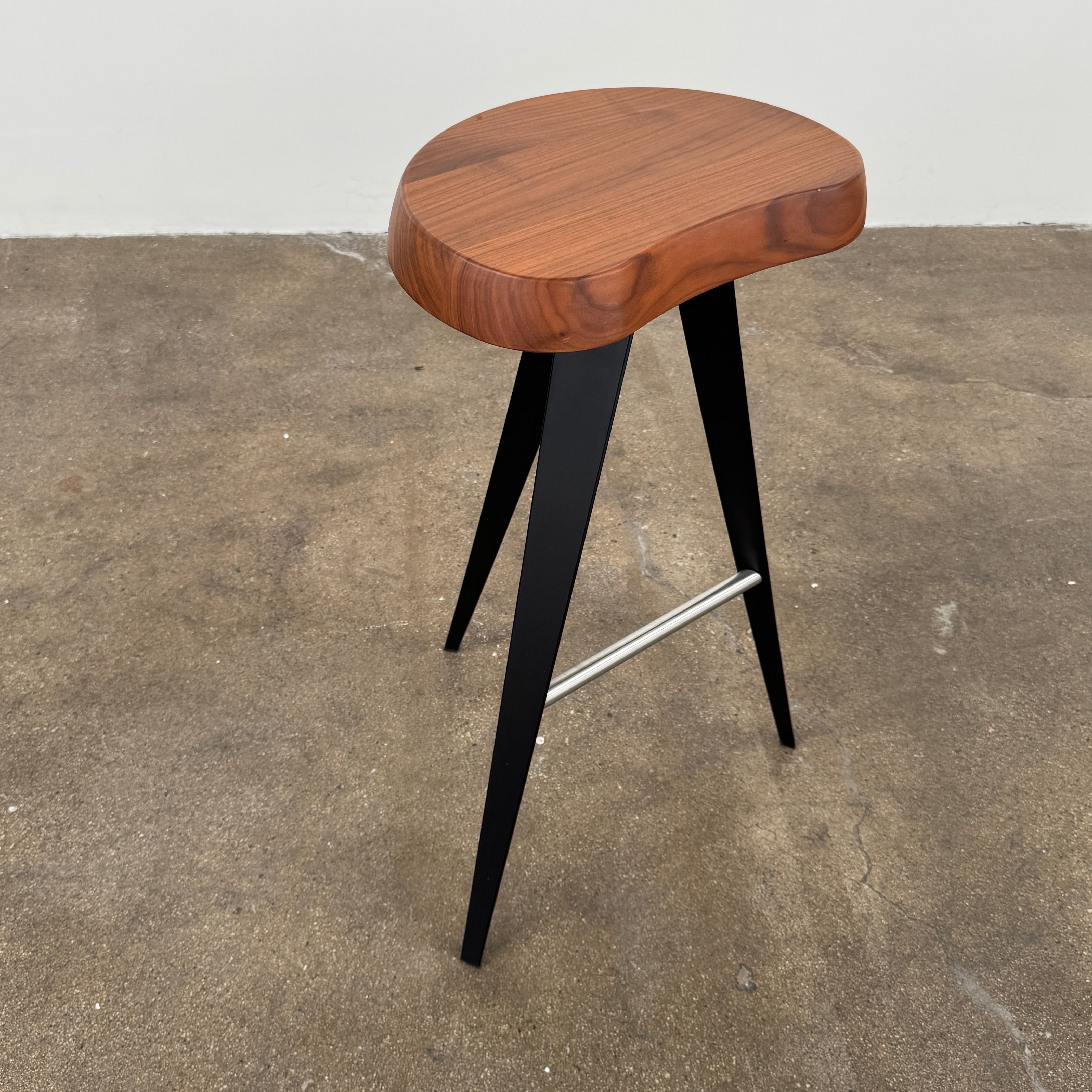 A Cassina Mexique Stool by Cassina, featuring a wooden round seat, three black angled legs, and a metal footrest, is displayed against a plain white background—highlighting its mid-century craftsmanship inspired by Charlotte Perriand.