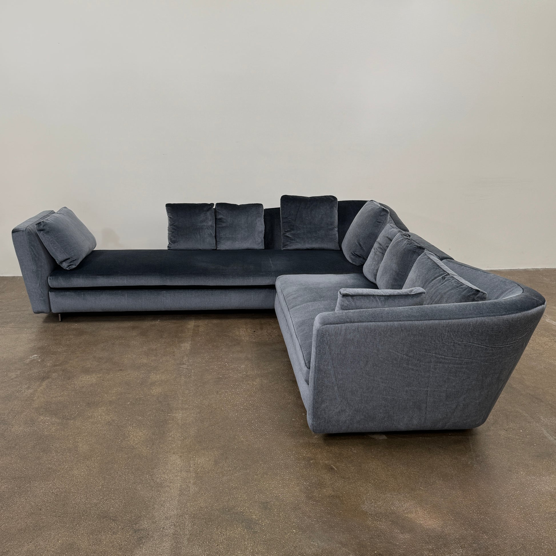 A Minotti Seymour Sofa by Modern Resale, featuring a gray L-shaped design with five back cushions, is situated on a brown concrete floor against a plain white wall.