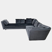 A contemporary Minotti Seymour Sofa, featuring a grey L-shaped design inspired by architectural forms, includes five matching back cushions and one side cushion, all upholstered in fabric and presented against a plain white background.