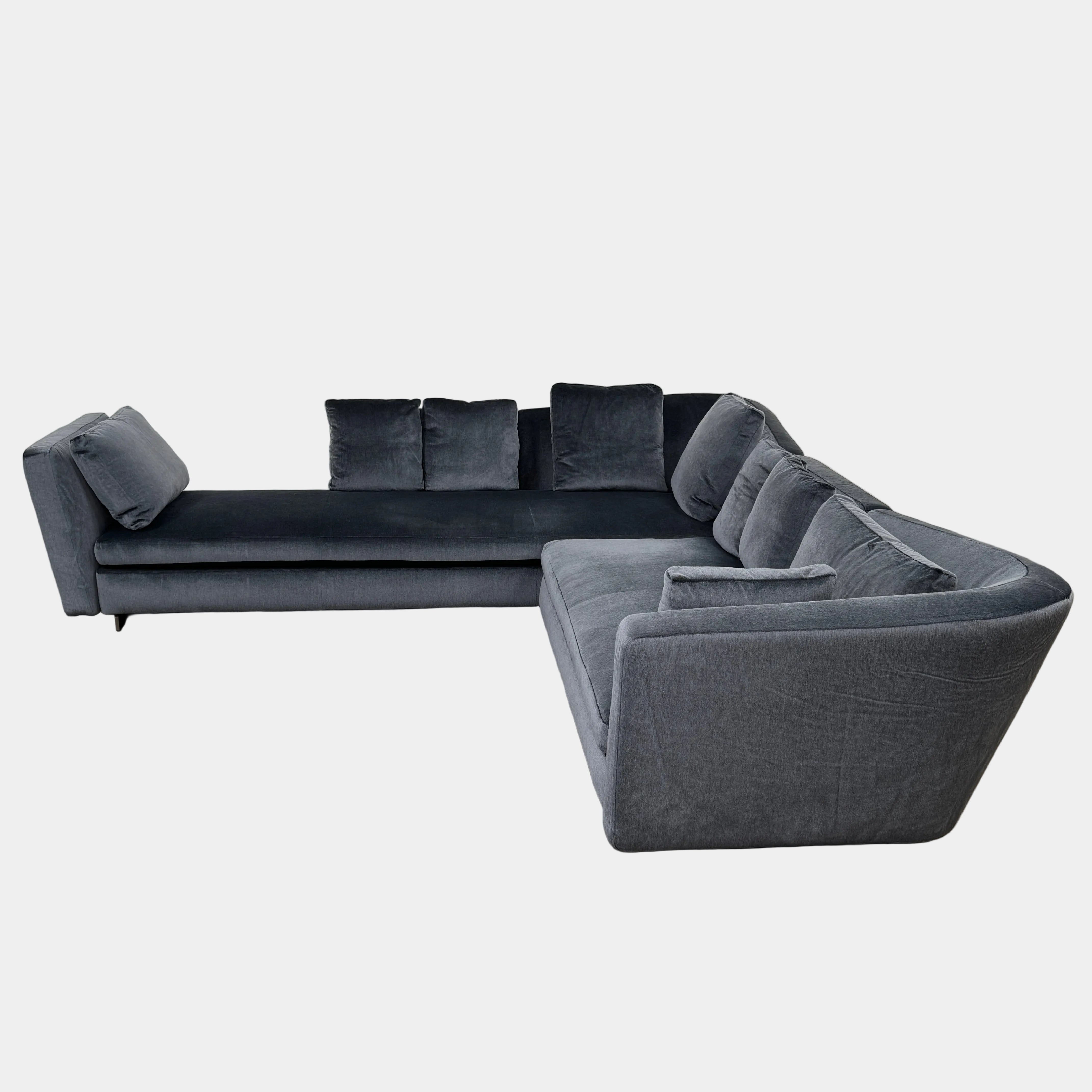 A contemporary Minotti Seymour Sofa, featuring a grey L-shaped design inspired by architectural forms, includes five matching back cushions and one side cushion, all upholstered in fabric and presented against a plain white background.