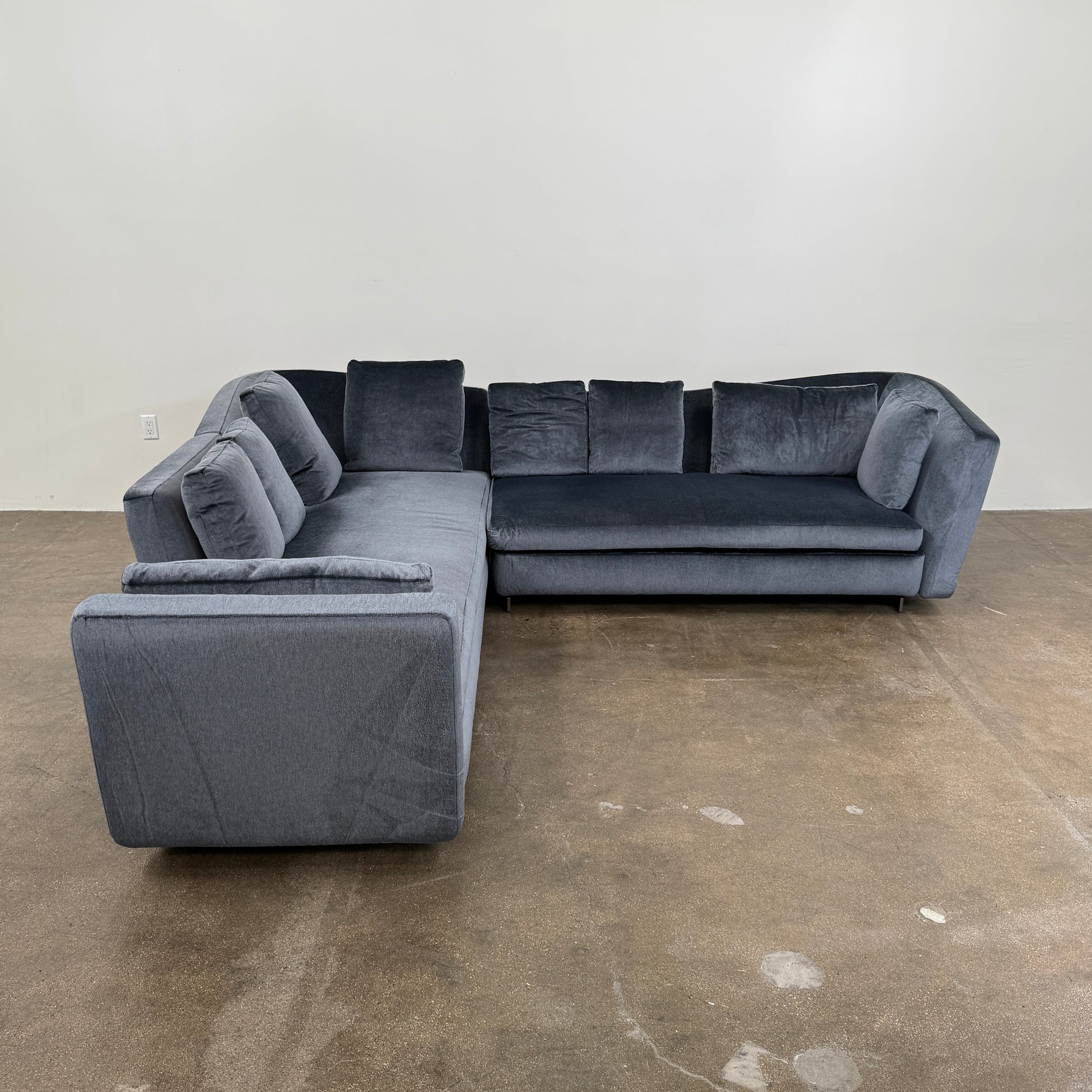 A contemporary Minotti Seymour Sofa, featuring a grey L-shaped design inspired by architectural forms, includes five matching back cushions and one side cushion, all upholstered in fabric and presented against a plain white background.