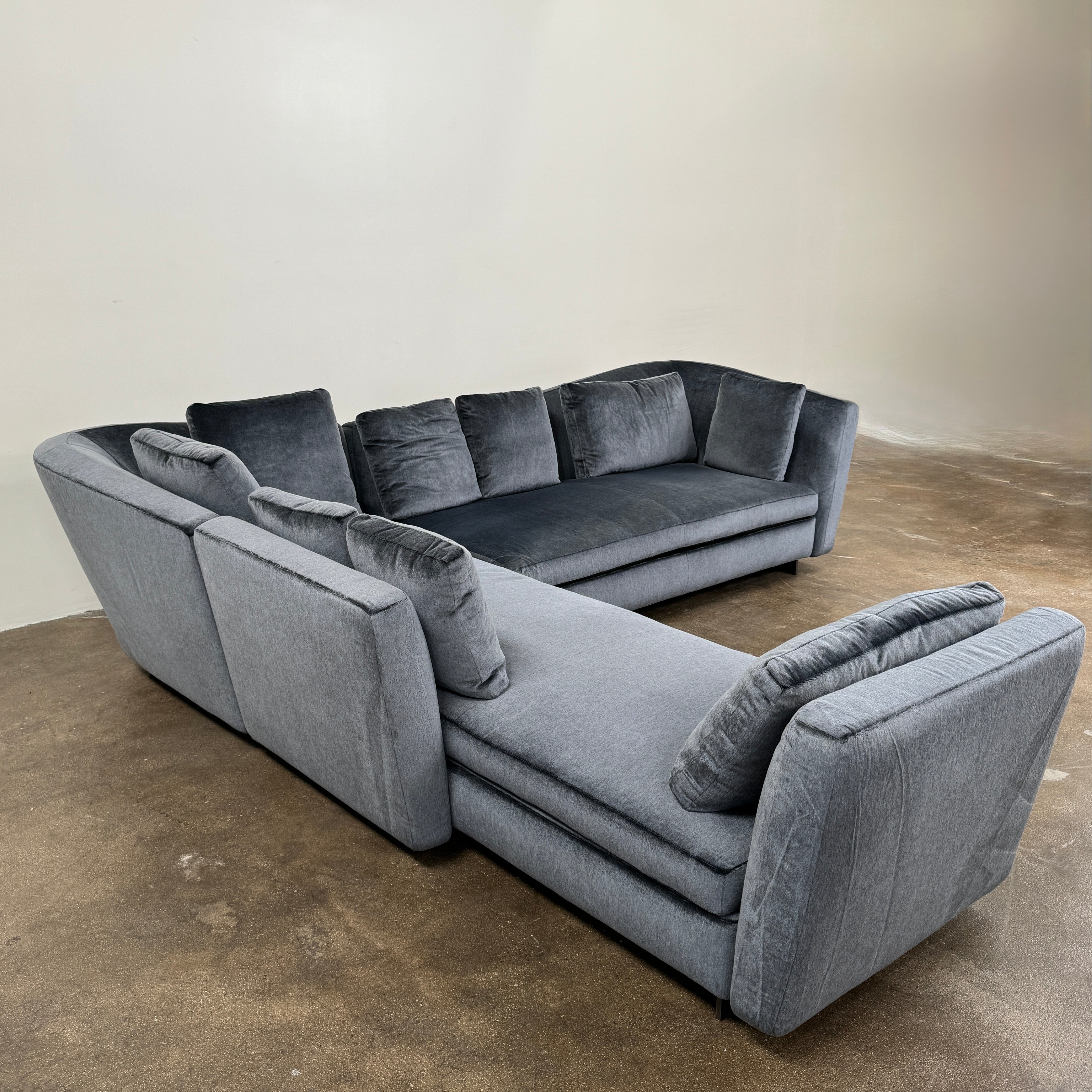 A contemporary Minotti Seymour Sofa, featuring a grey L-shaped design inspired by architectural forms, includes five matching back cushions and one side cushion, all upholstered in fabric and presented against a plain white background.