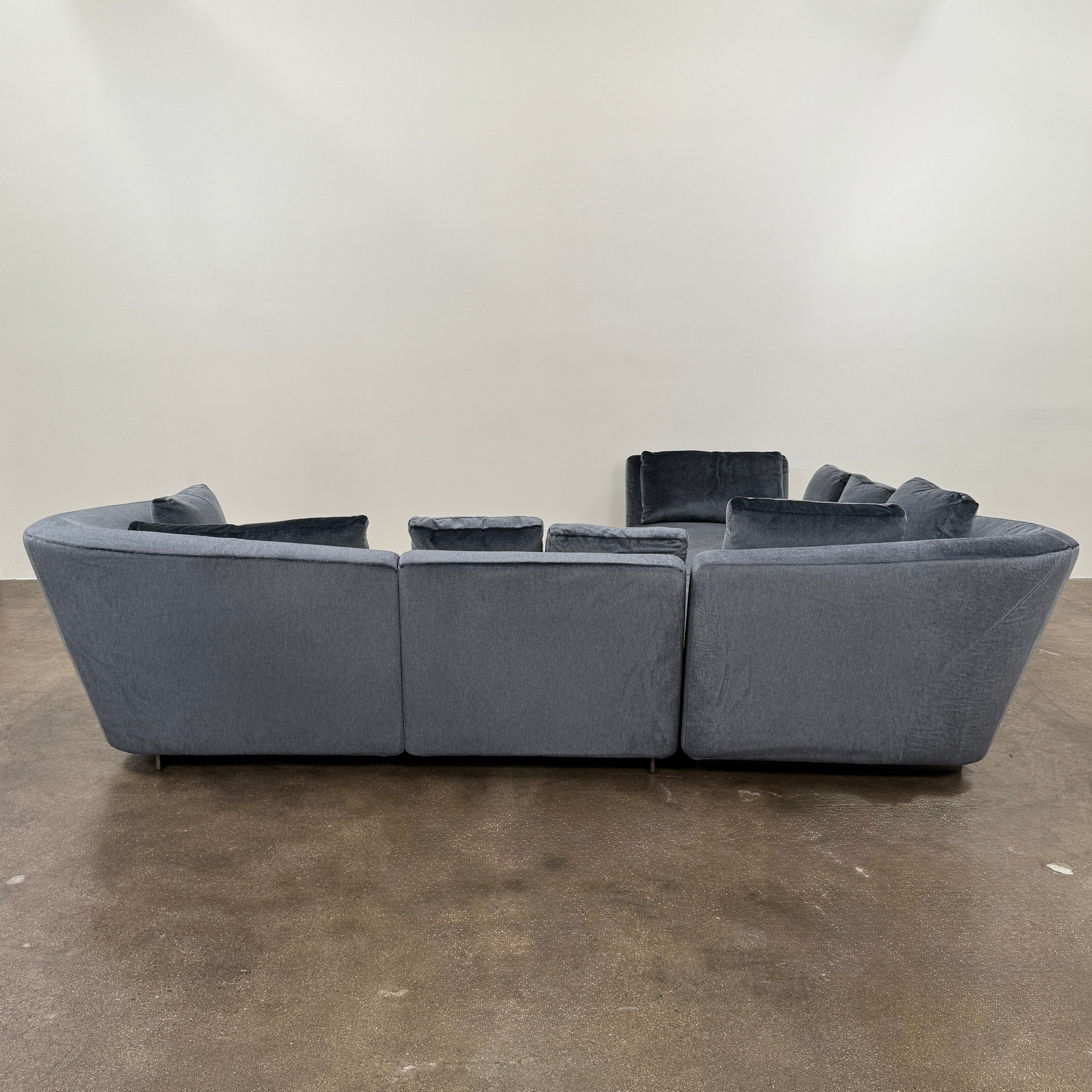 A contemporary Minotti Seymour Sofa, featuring a grey L-shaped design inspired by architectural forms, includes five matching back cushions and one side cushion, all upholstered in fabric and presented against a plain white background.