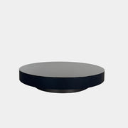 A large, round Minotti Milton Coffee Table in black rests on a polished concrete floor against a plain white wall, evoking the aesthetic of contemporary design.