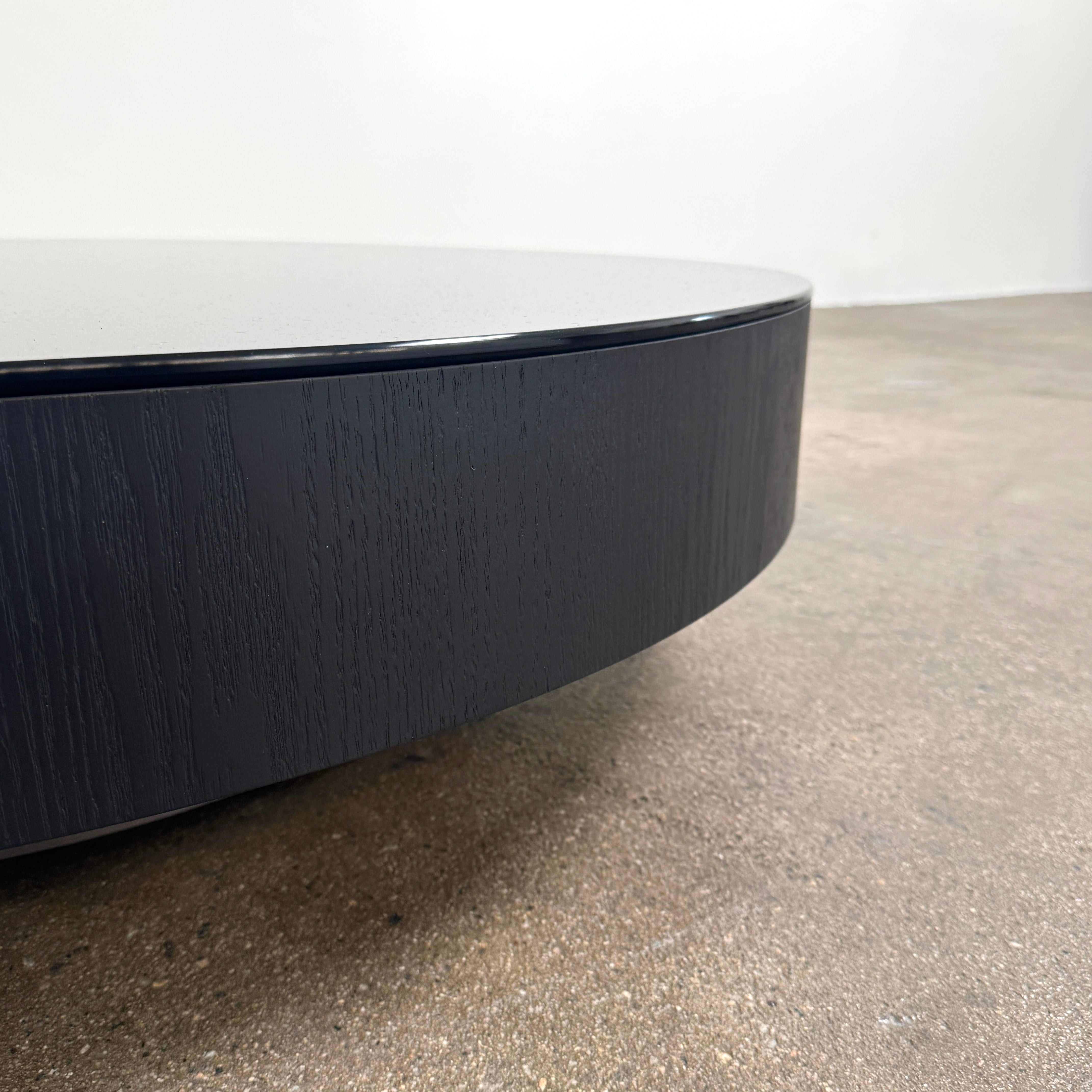A large, round Minotti Milton Coffee Table in black rests on a polished concrete floor against a plain white wall, evoking the aesthetic of contemporary design.