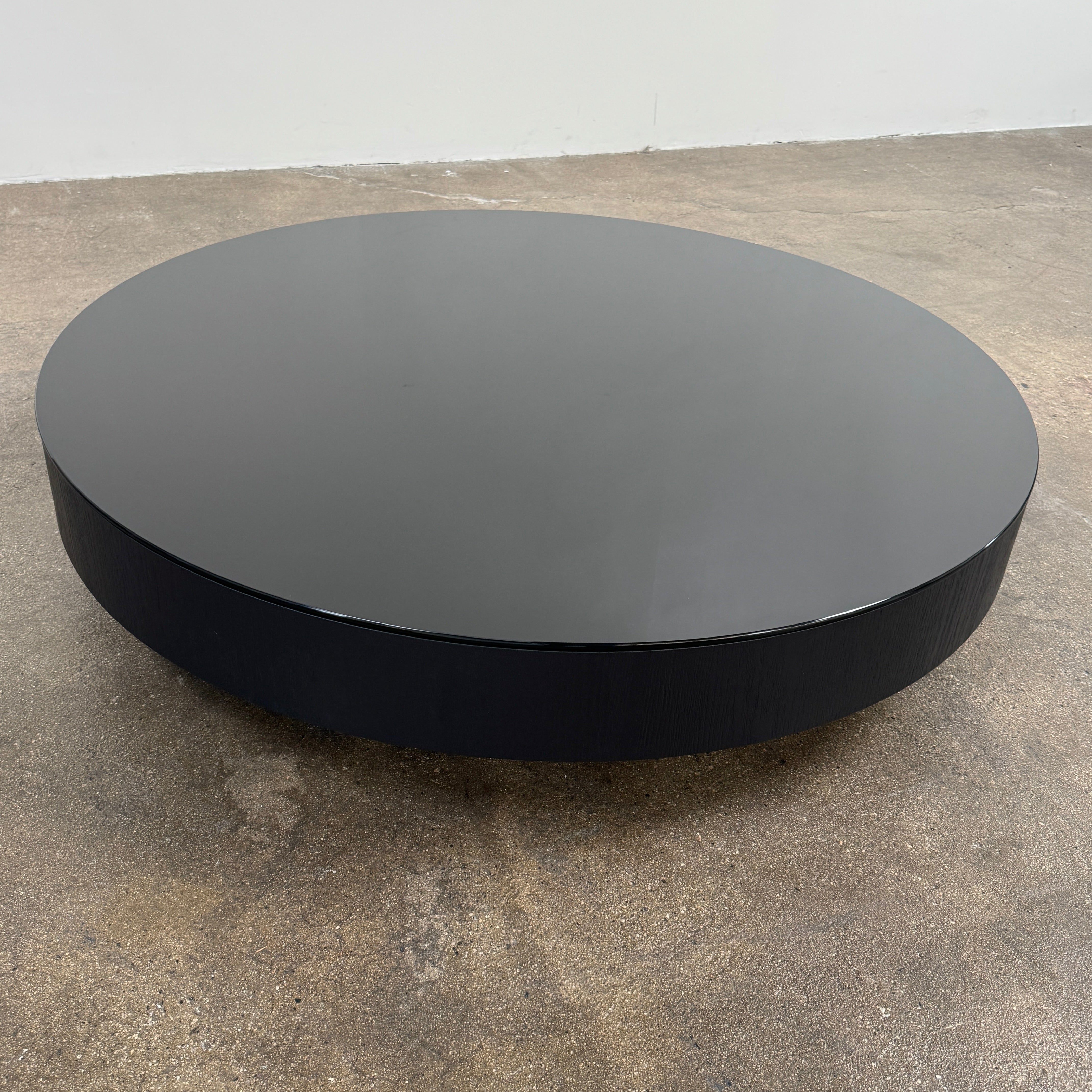 A large, round Minotti Milton Coffee Table in black rests on a polished concrete floor against a plain white wall, evoking the aesthetic of contemporary design.