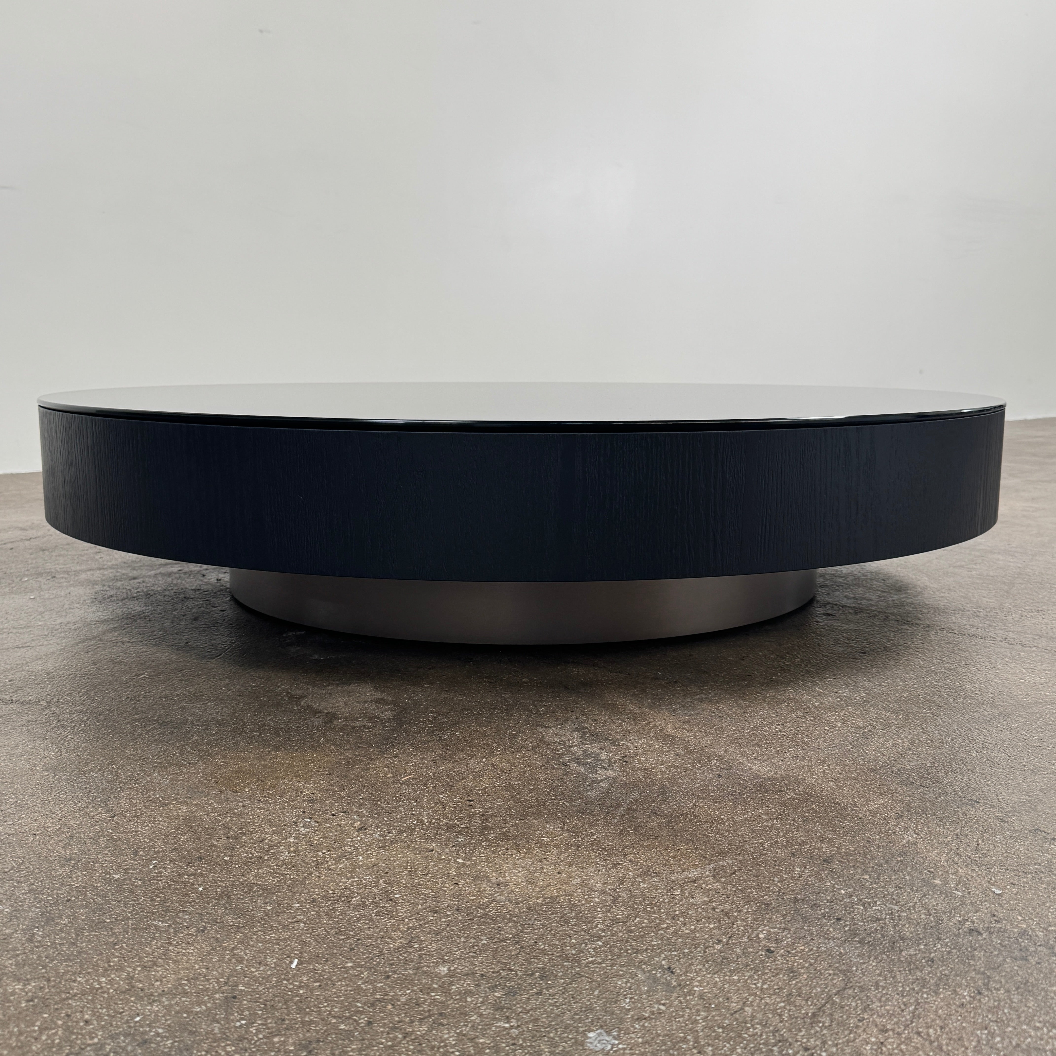 A large, round Minotti Milton Coffee Table in black rests on a polished concrete floor against a plain white wall, evoking the aesthetic of contemporary design.