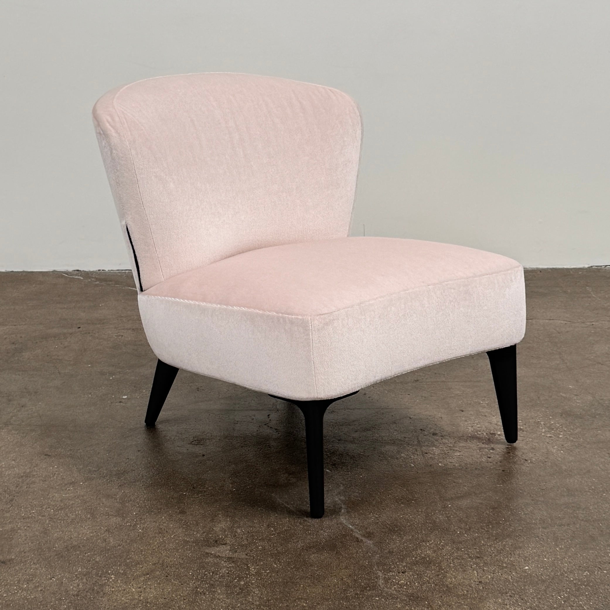 The Minotti Aston armless Armchair by Modern Resale, featuring light pink reflective upholstery and dark wooden legs, is placed on a concrete floor against a plain light gray wall.