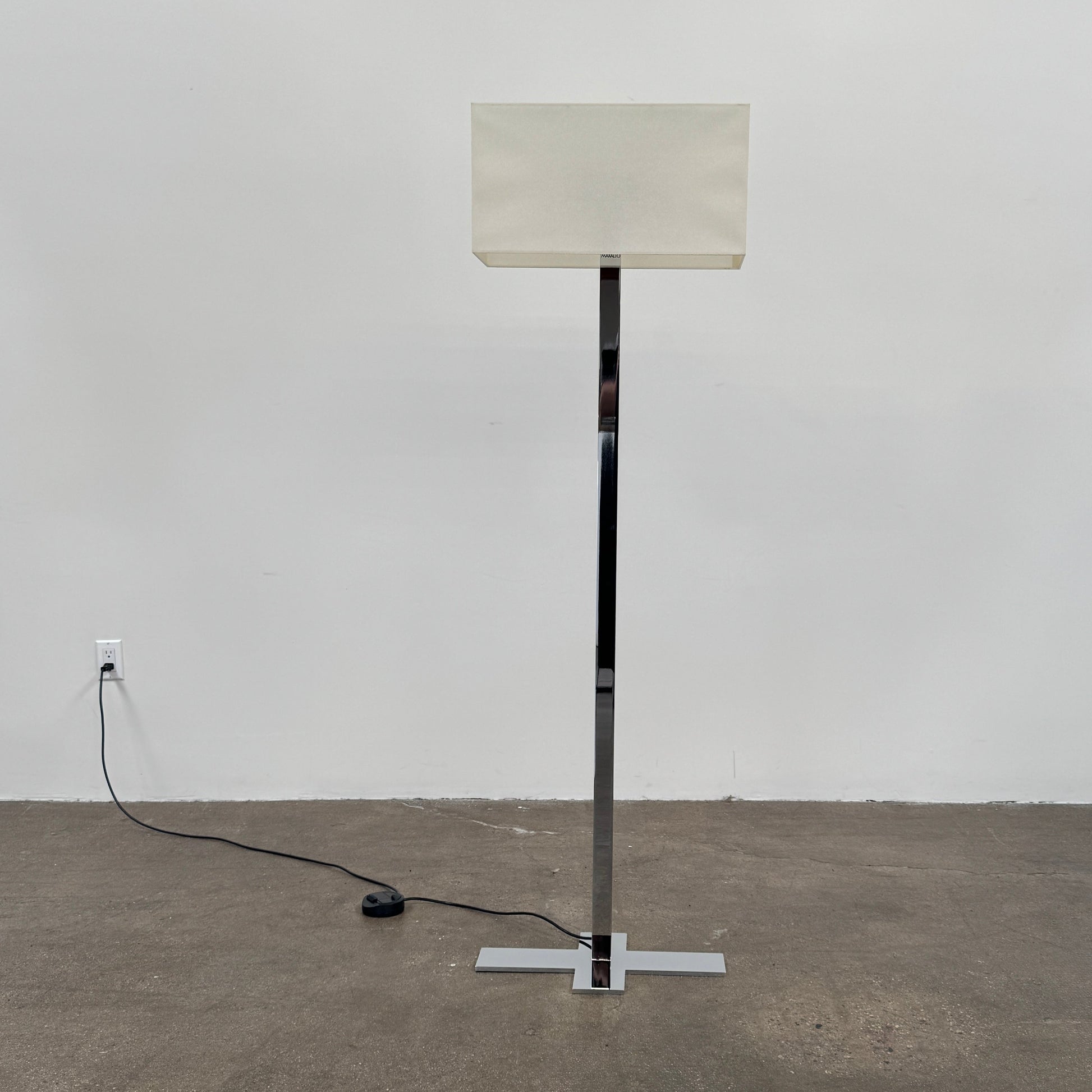 The Maxalto Leukon Floor Lamp by Maxalto features a tall chrome stand, rectangular white shade, distinctive cross-shaped base, and a black power cord with foot switch, set against a plain background.