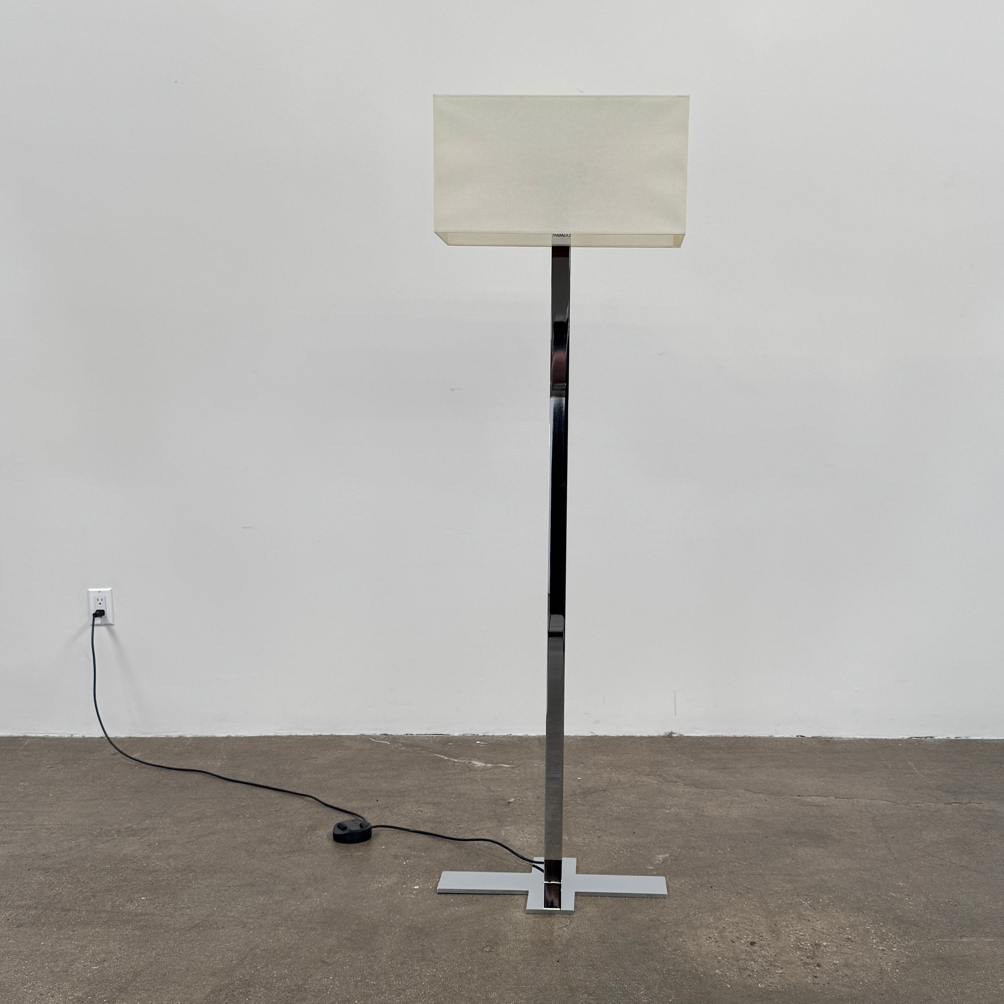 The Maxalto Leukon Floor Lamp by Maxalto features a tall chrome stand, rectangular white shade, distinctive cross-shaped base, and a black power cord with foot switch, set against a plain background.