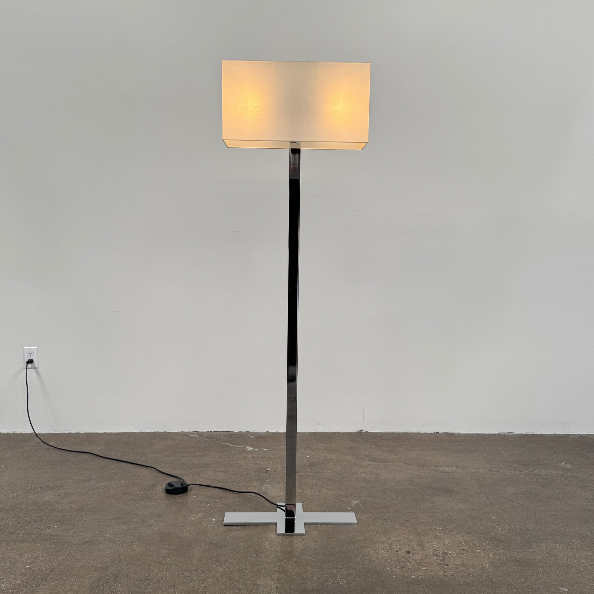 The Maxalto Leukon Floor Lamp by Maxalto features a tall chrome stand, rectangular white shade, distinctive cross-shaped base, and a black power cord with foot switch, set against a plain background.