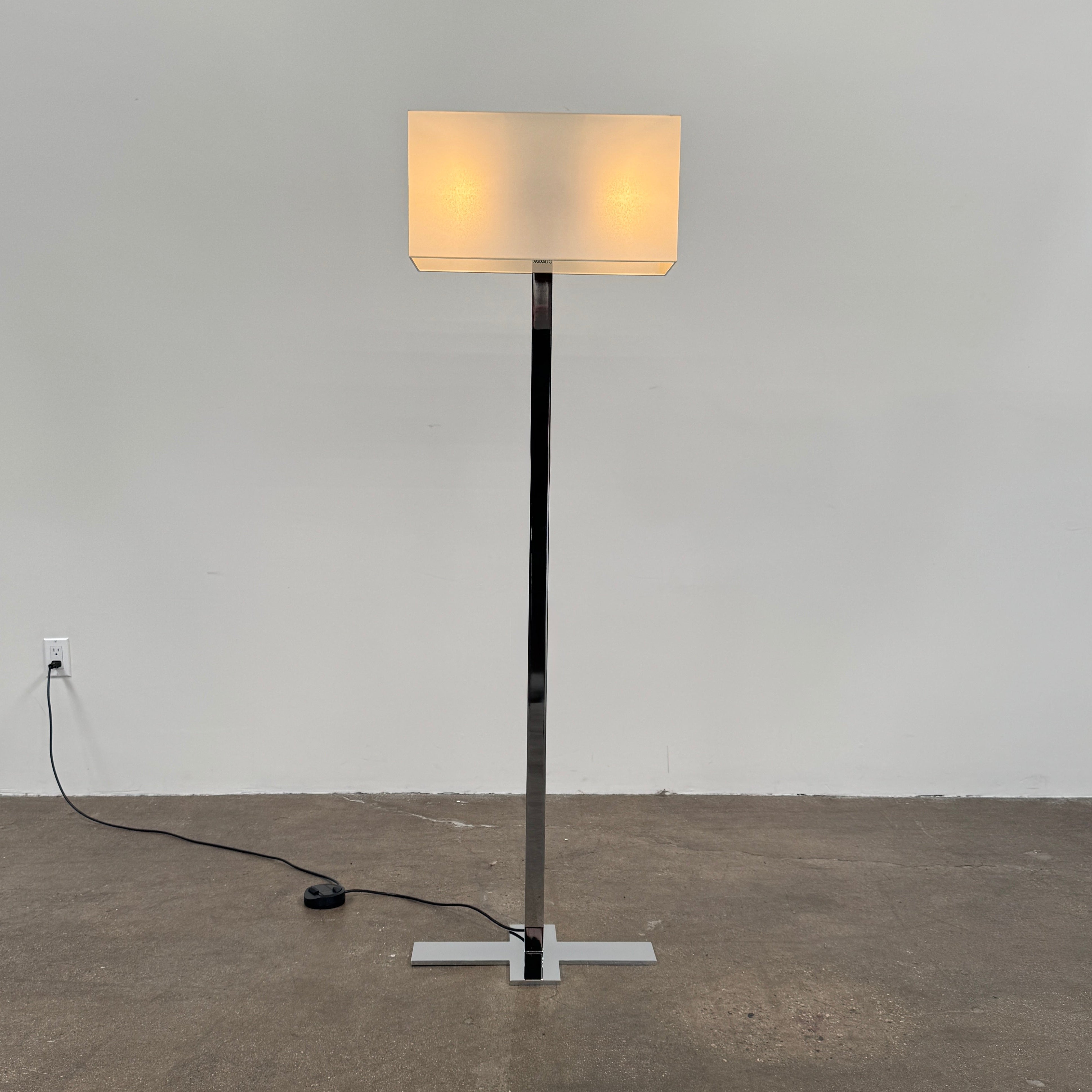 The Maxalto Leukon Floor Lamp by Maxalto features a tall chrome stand, rectangular white shade, distinctive cross-shaped base, and a black power cord with foot switch, set against a plain background.