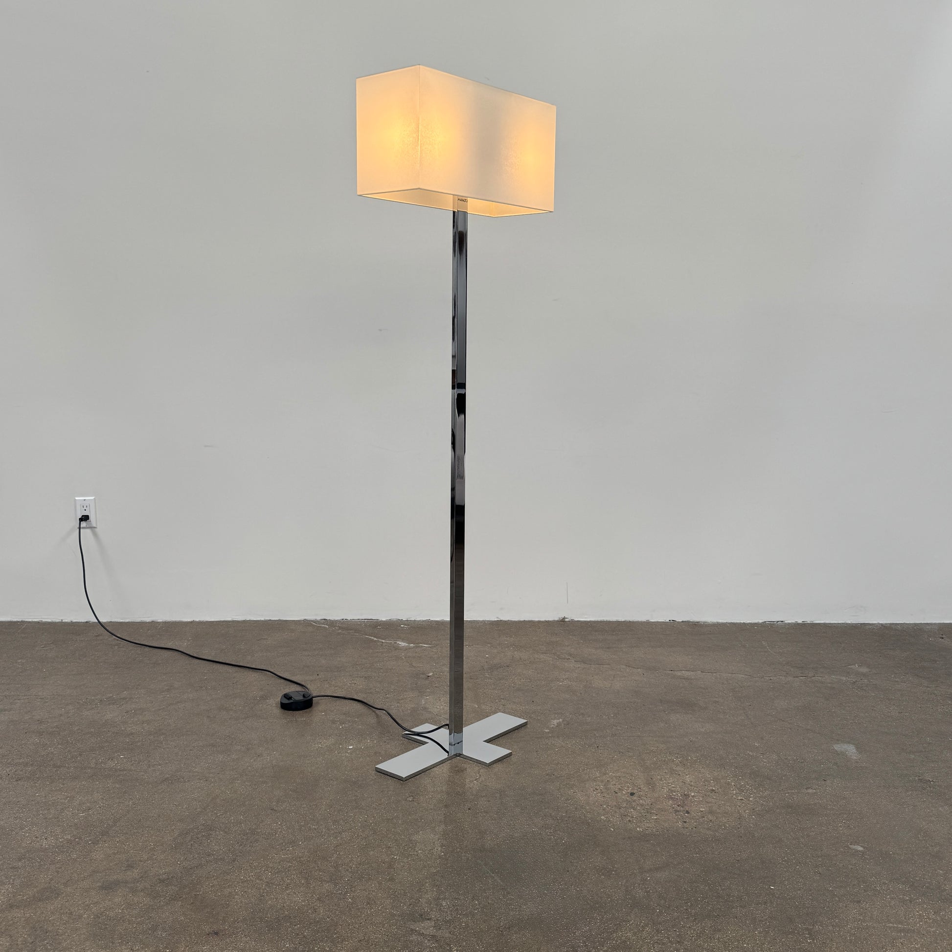 The Maxalto Leukon Floor Lamp by Maxalto features a tall chrome stand, rectangular white shade, distinctive cross-shaped base, and a black power cord with foot switch, set against a plain background.