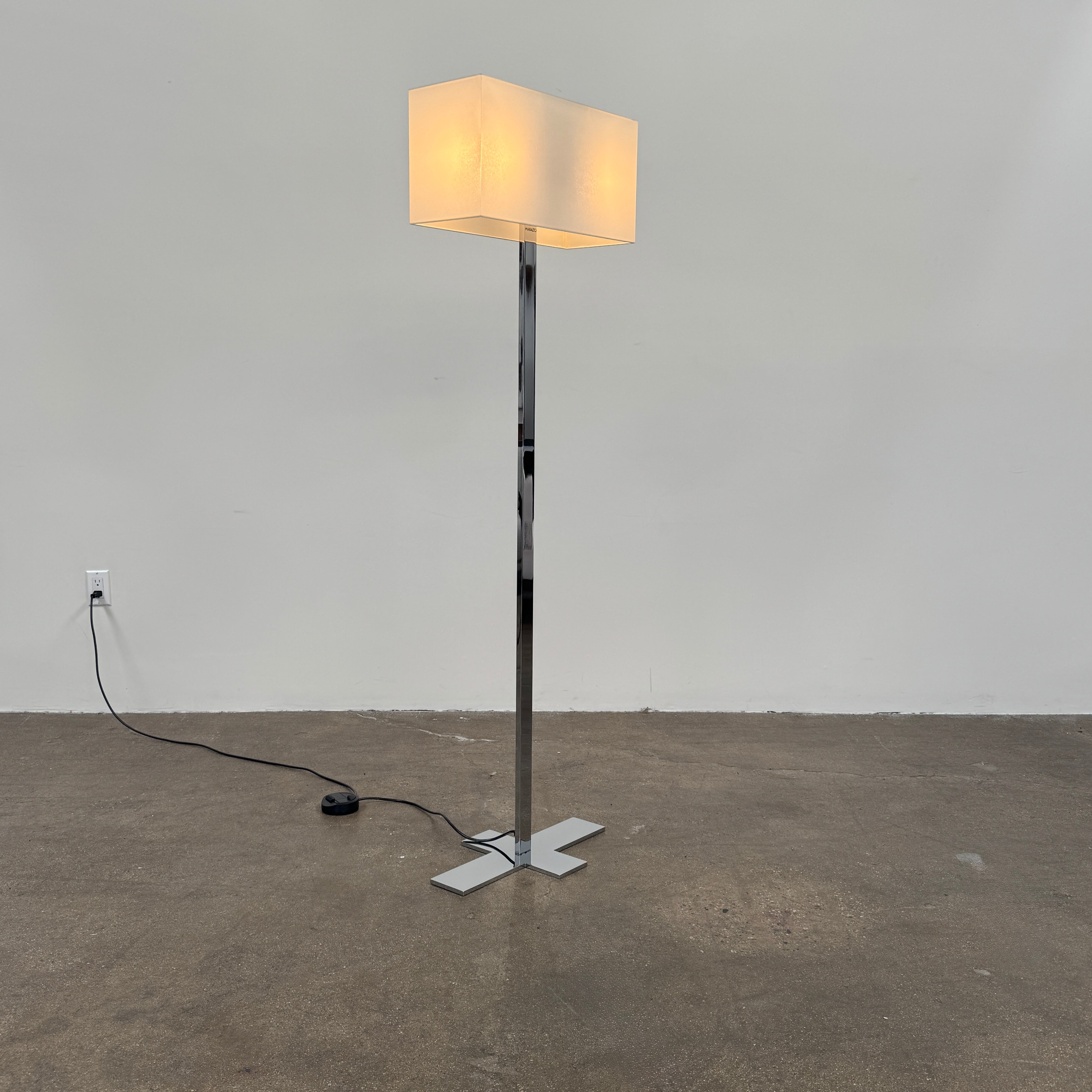 The Maxalto Leukon Floor Lamp by Maxalto features a tall chrome stand, rectangular white shade, distinctive cross-shaped base, and a black power cord with foot switch, set against a plain background.