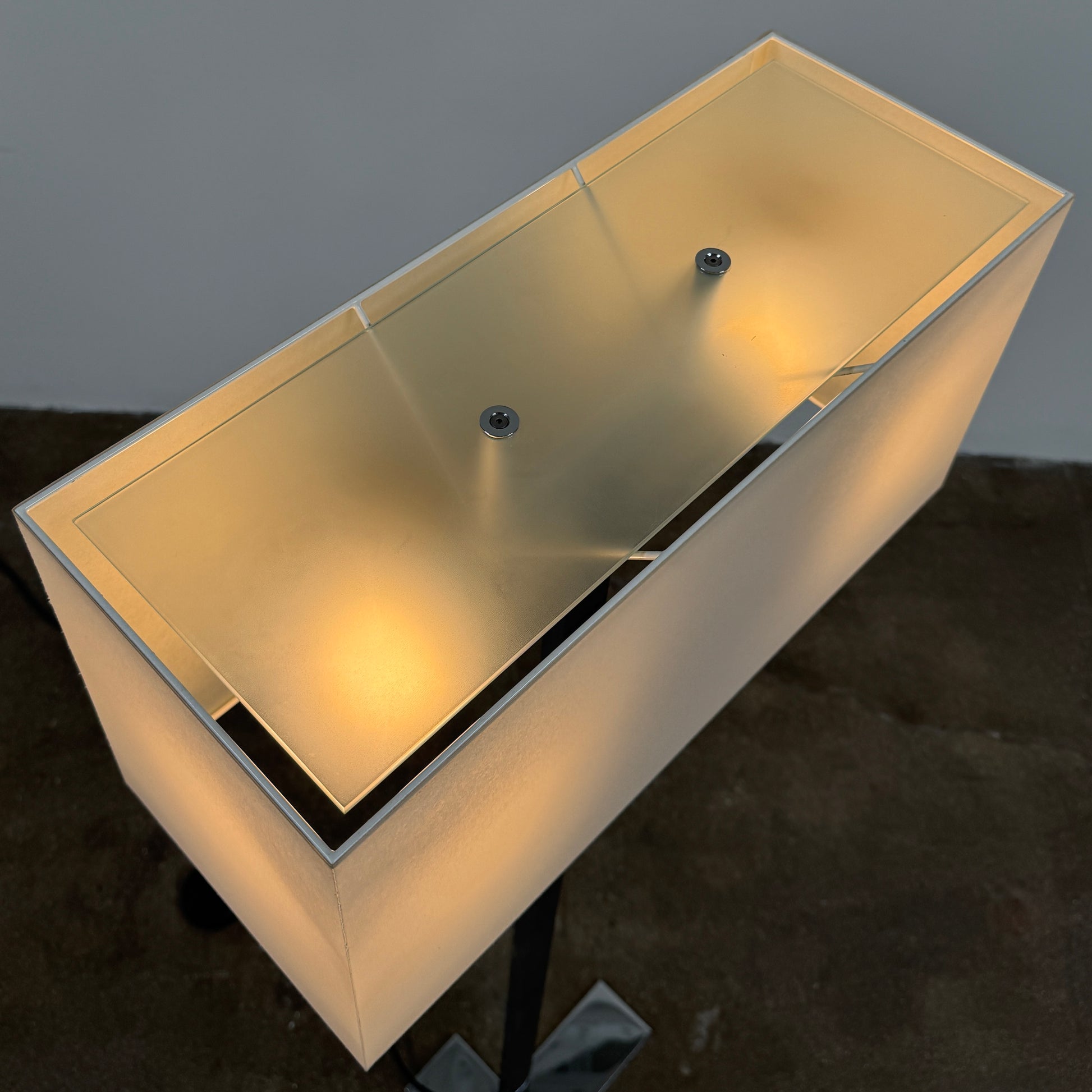 The Maxalto Leukon Floor Lamp by Maxalto features a tall chrome stand, rectangular white shade, distinctive cross-shaped base, and a black power cord with foot switch, set against a plain background.