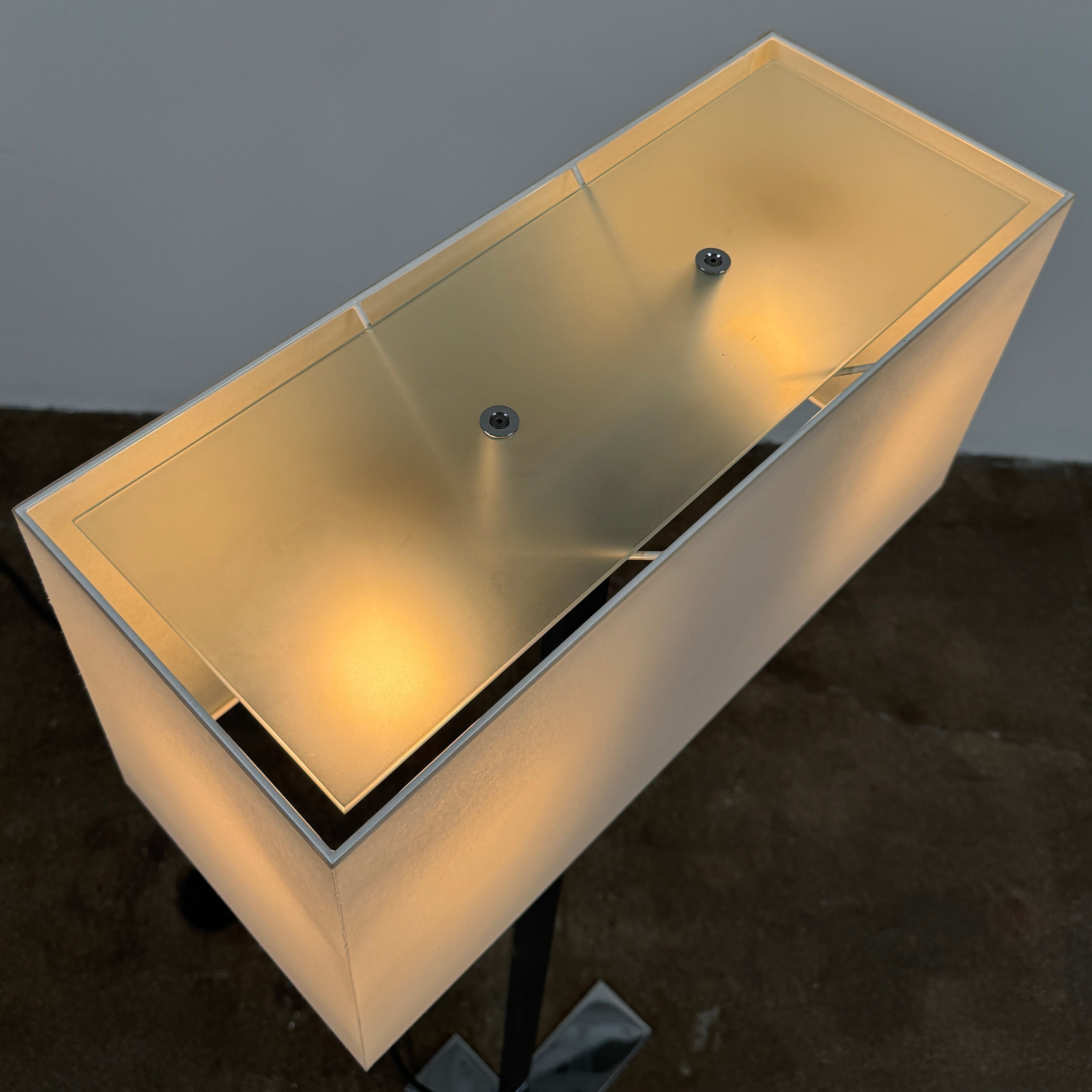 The Maxalto Leukon Floor Lamp by Maxalto features a tall chrome stand, rectangular white shade, distinctive cross-shaped base, and a black power cord with foot switch, set against a plain background.