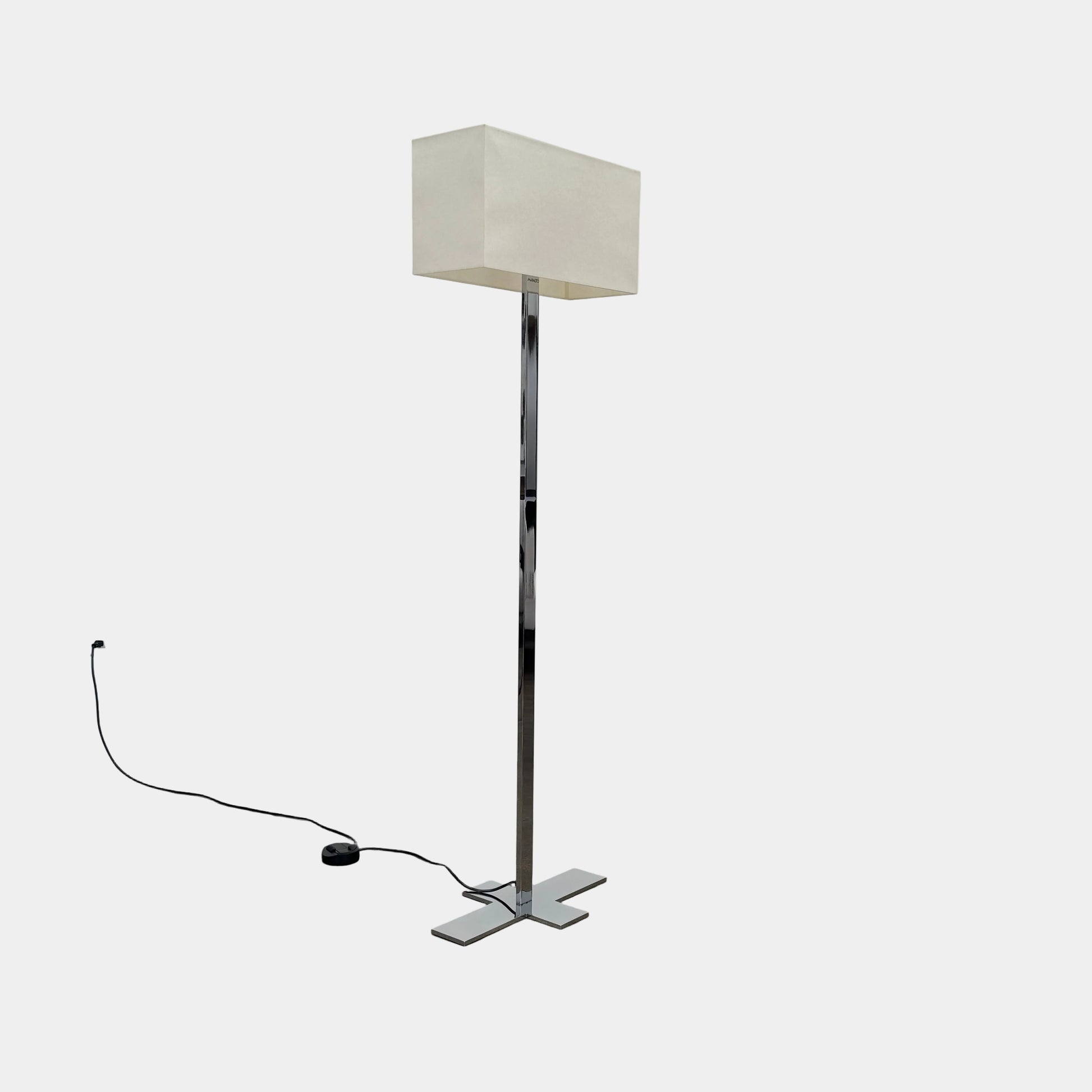 The Maxalto Leukon Floor Lamp by Maxalto features a tall chrome stand, rectangular white shade, distinctive cross-shaped base, and a black power cord with foot switch, set against a plain background.