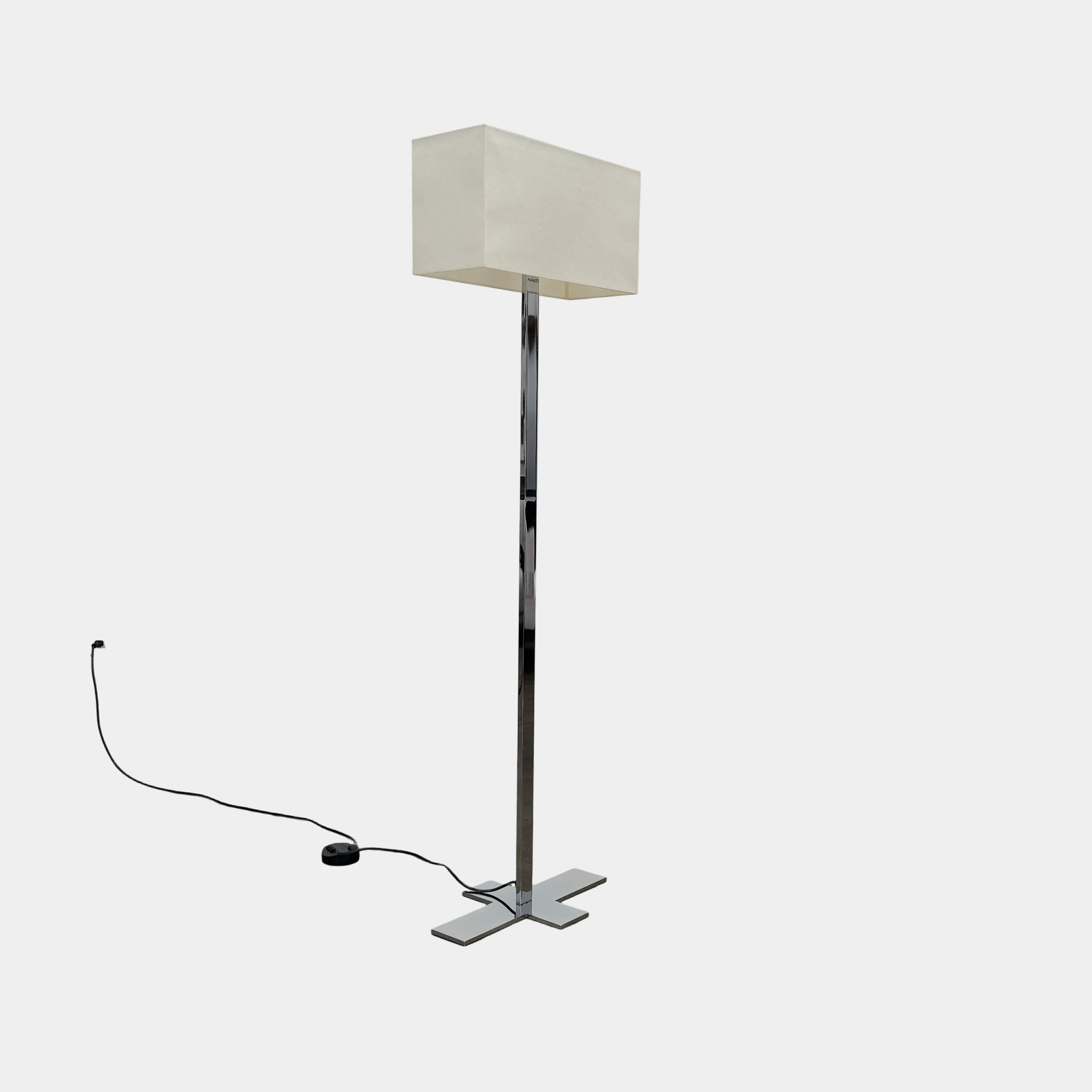 The Maxalto Leukon Floor Lamp by Maxalto features a tall chrome stand, rectangular white shade, distinctive cross-shaped base, and a black power cord with foot switch, set against a plain background.