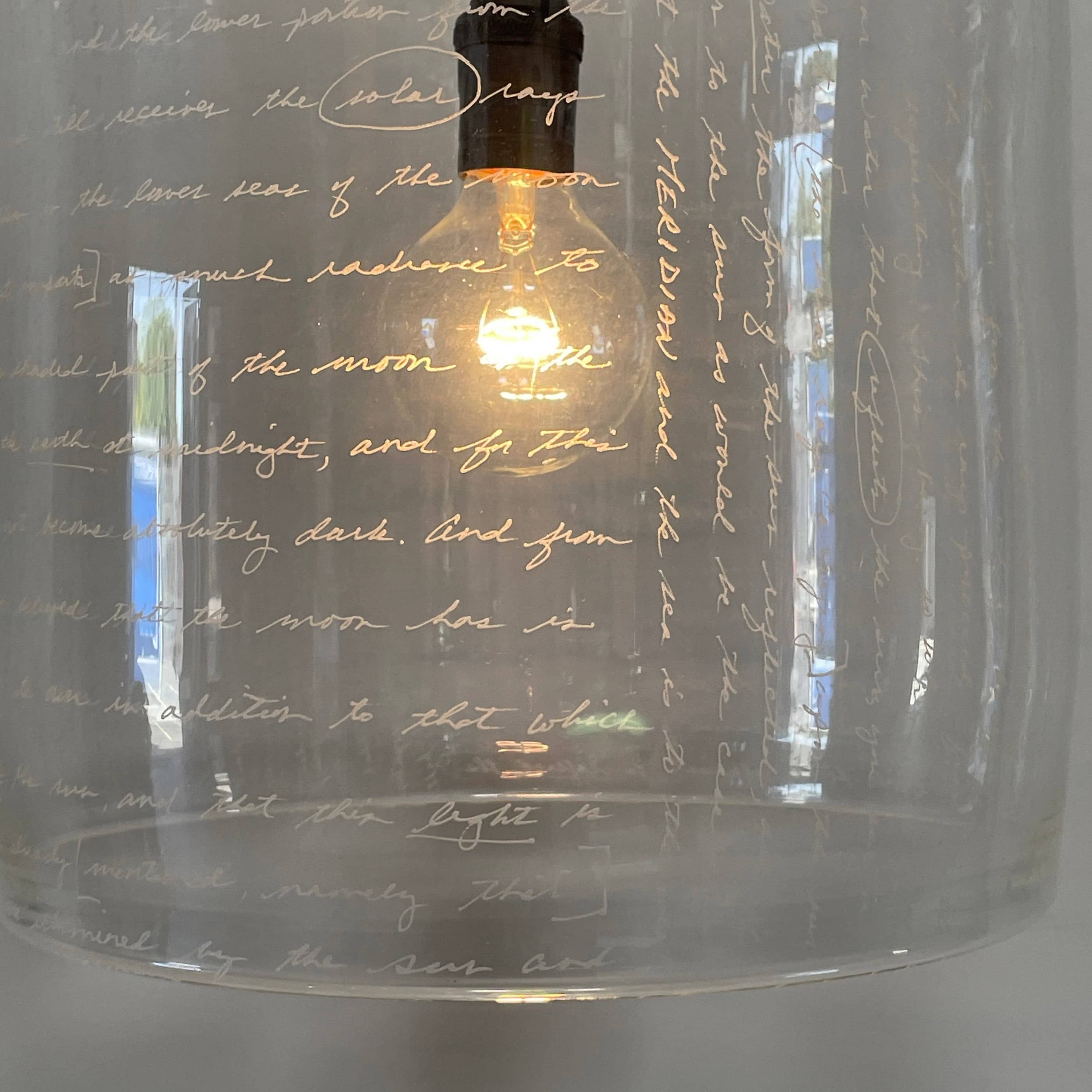 A unique functional art piece, this Alison Berger Word Pendant by Alison Berger showcases visible wires and mounting hardware against a white textured ceiling, reminiscent of Alison Berger's Word Pendant designs.