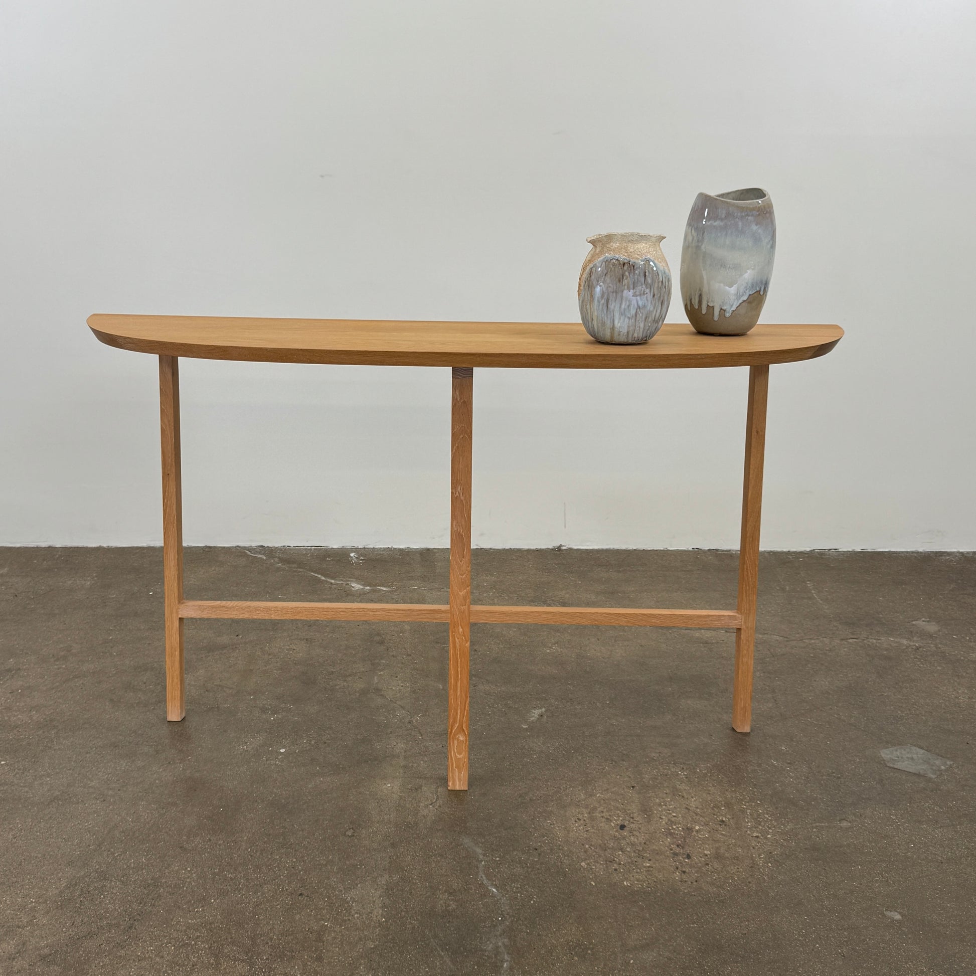 A De La Espada Trio Console Table 754CL by De La Espada stands against a white wall, showcasing modern woodworking with three vases, a beaded necklace, and a small stack of books arranged on its surface atop the concrete floor.