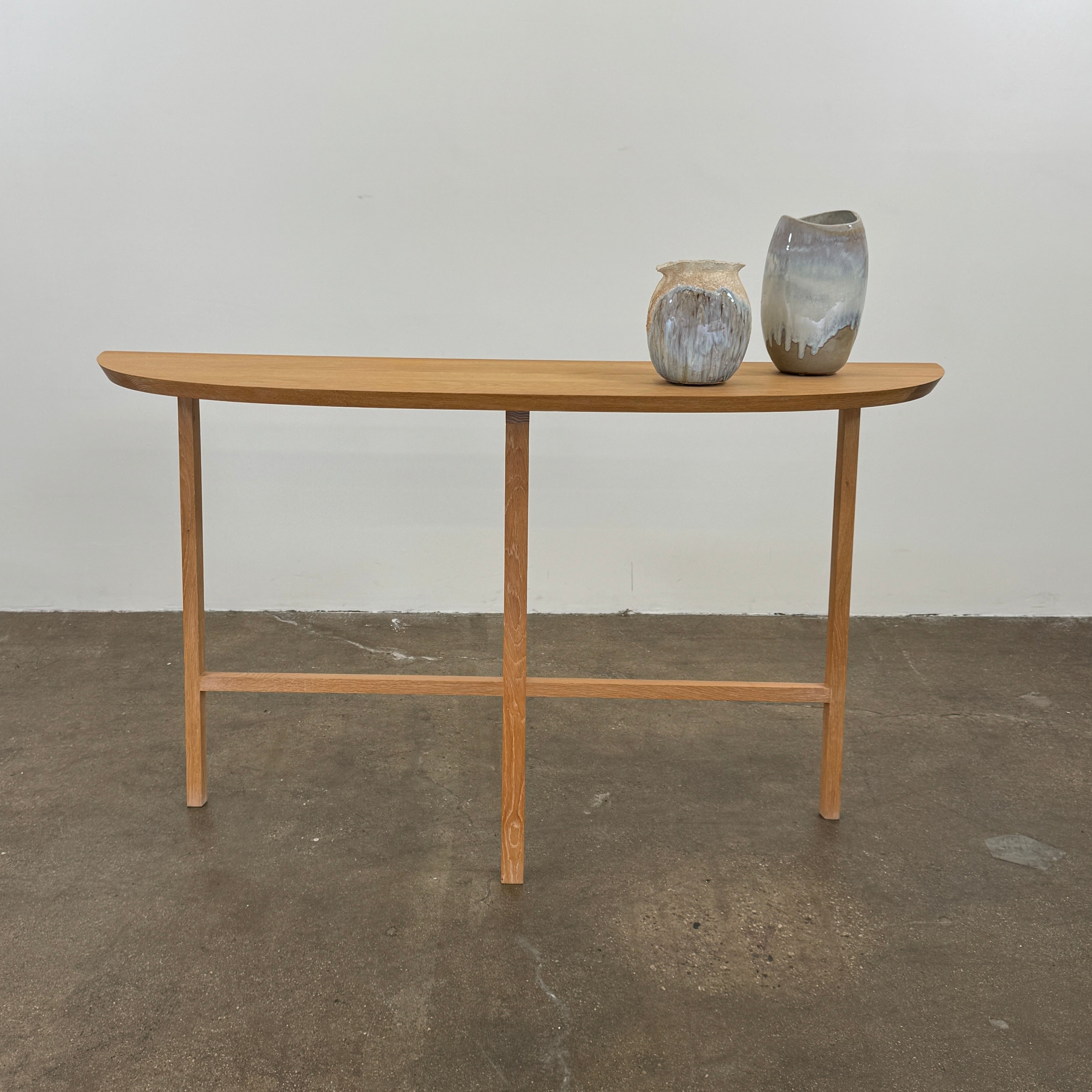 A De La Espada Trio Console Table 754CL by De La Espada stands against a white wall, showcasing modern woodworking with three vases, a beaded necklace, and a small stack of books arranged on its surface atop the concrete floor.