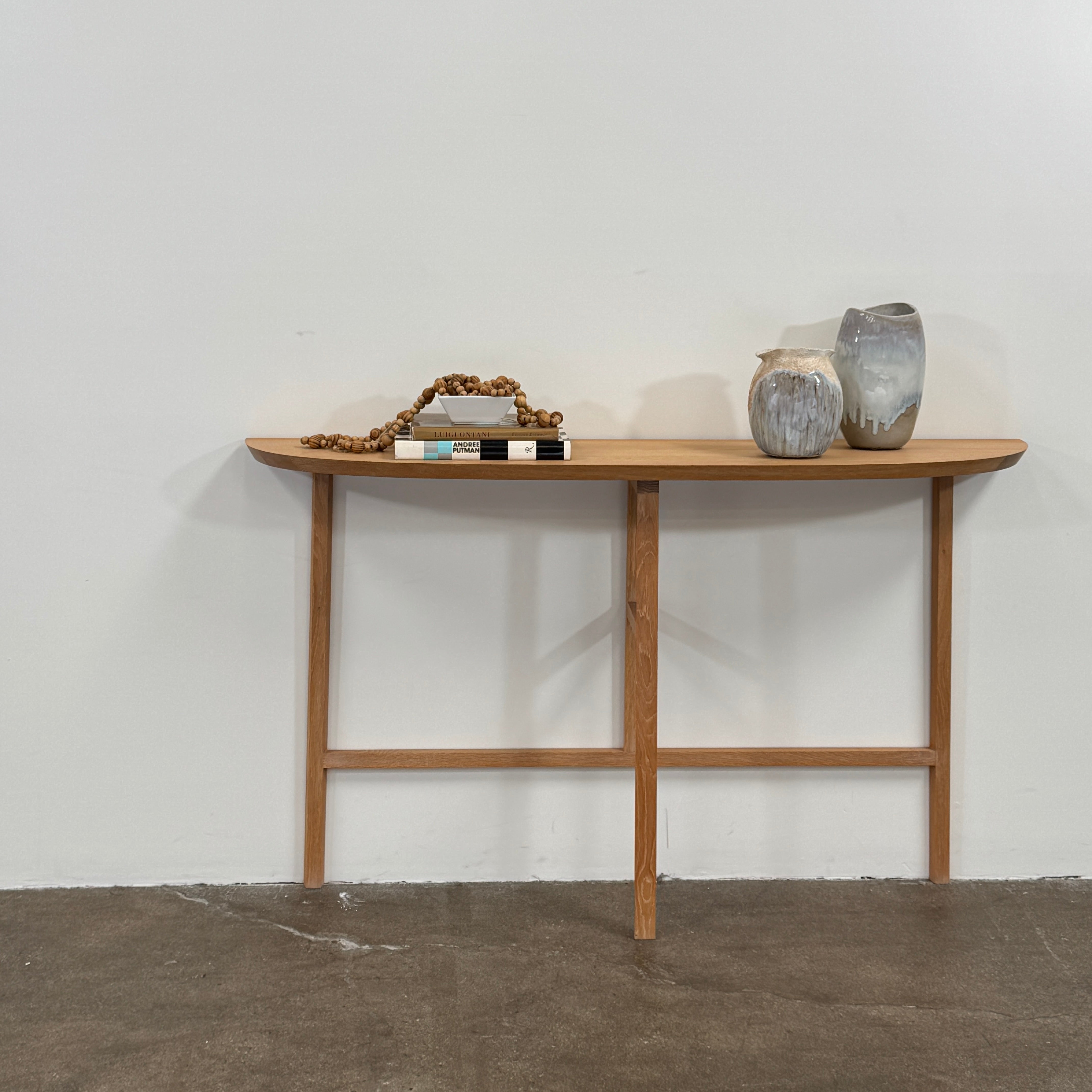 A De La Espada Trio Console Table 754CL by De La Espada stands against a white wall, showcasing modern woodworking with three vases, a beaded necklace, and a small stack of books arranged on its surface atop the concrete floor.