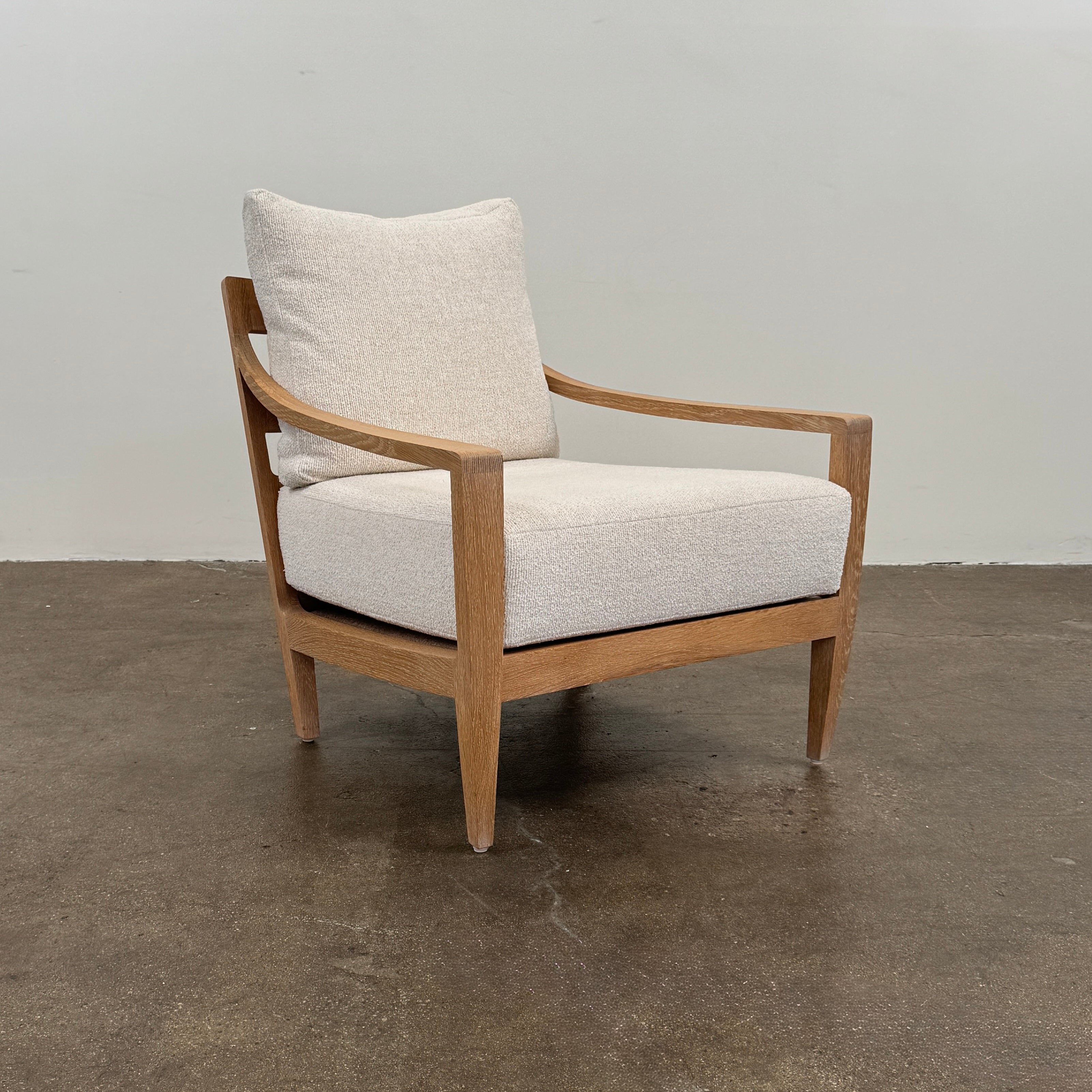 The De La Espada Low Lounge Chair 340 with Boucle material, from Modern Resale, sits on a polished concrete floor against a plain light-colored wall. Its wooden frame and light beige cushions evoke designer furniture and reflect the style of Matthew Hilton.