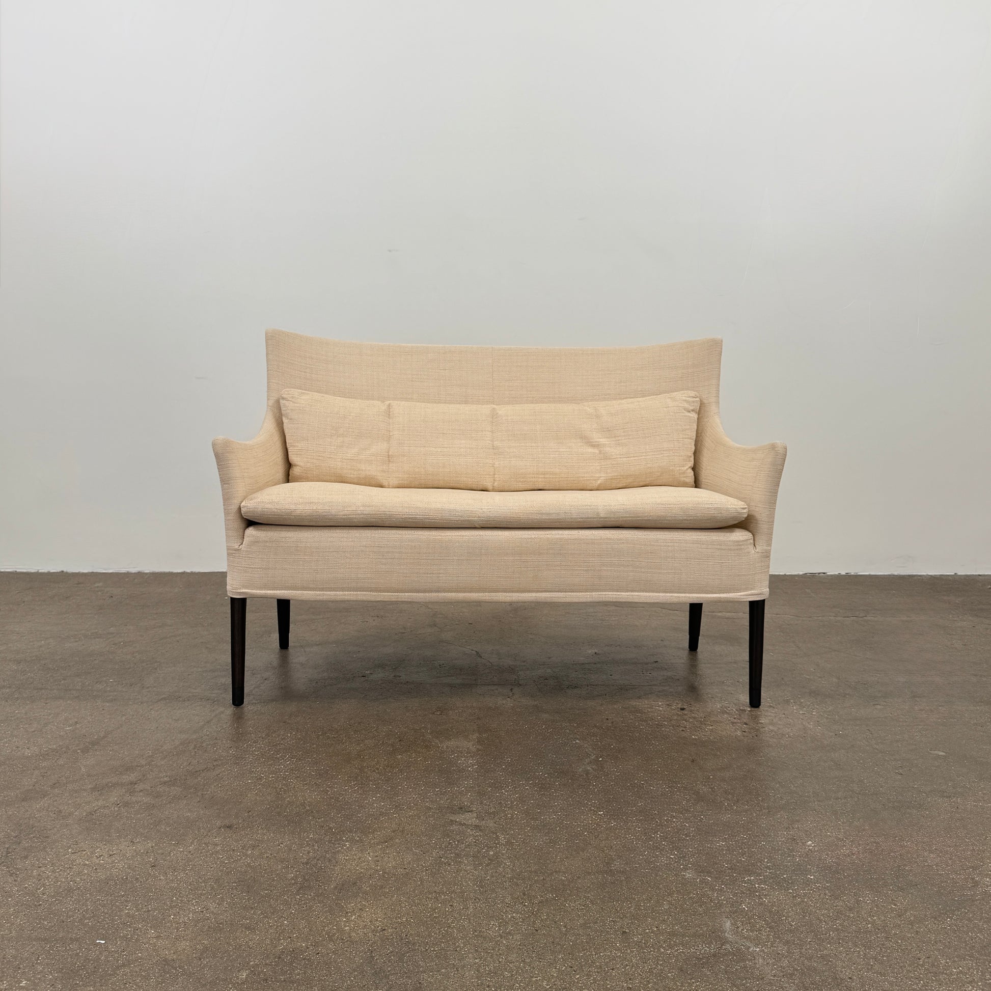 A Vintage Giorgetti Settee by Giorgetti, upholstered in beige with two matching cushions, black legs, and a high back, is placed on a polished concrete floor against a plain white wall.