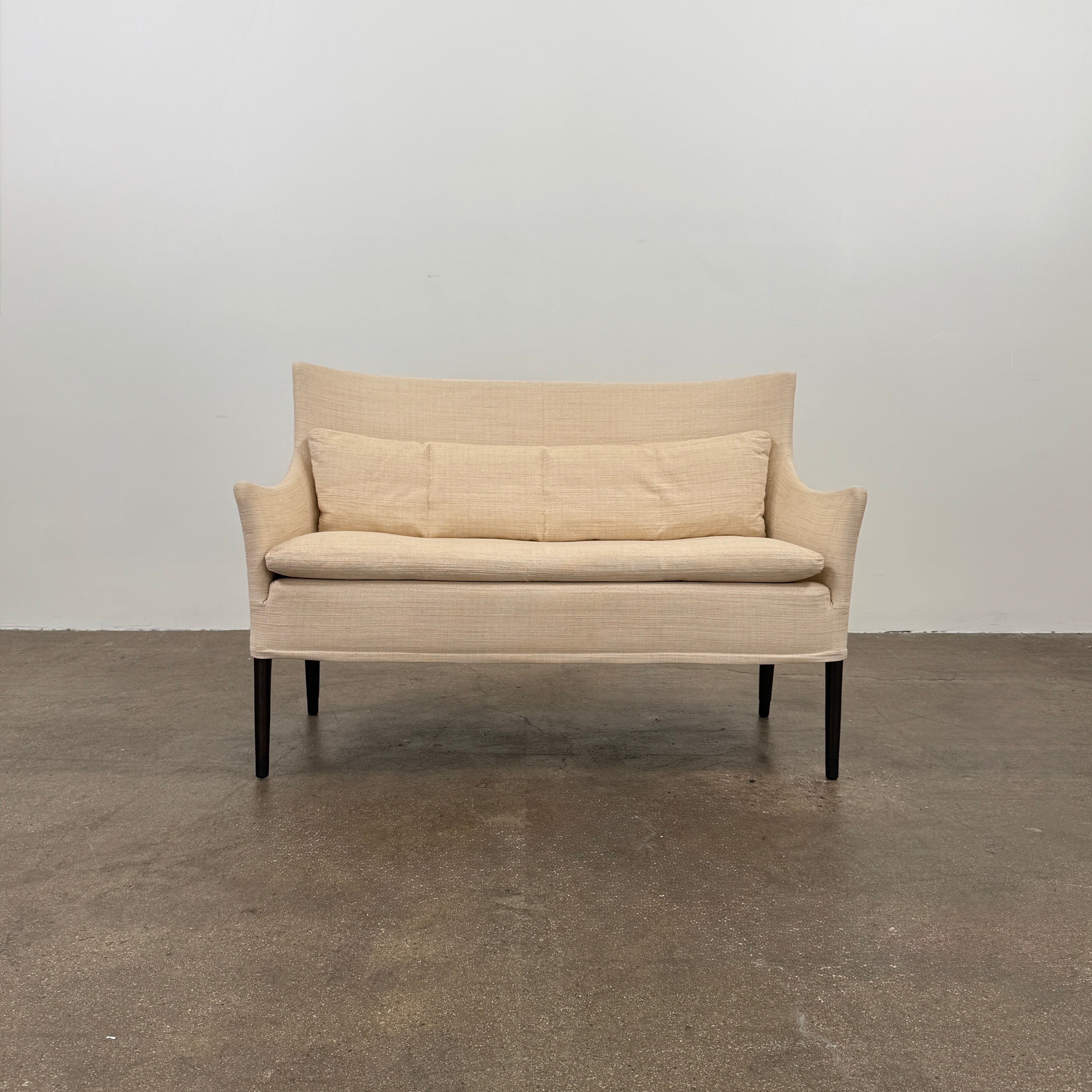 A Vintage Giorgetti Settee by Giorgetti, upholstered in beige with two matching cushions, black legs, and a high back, is placed on a polished concrete floor against a plain white wall.
