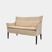 A Vintage Giorgetti Settee by Giorgetti, upholstered in beige with two matching cushions, black legs, and a high back, is placed on a polished concrete floor against a plain white wall.