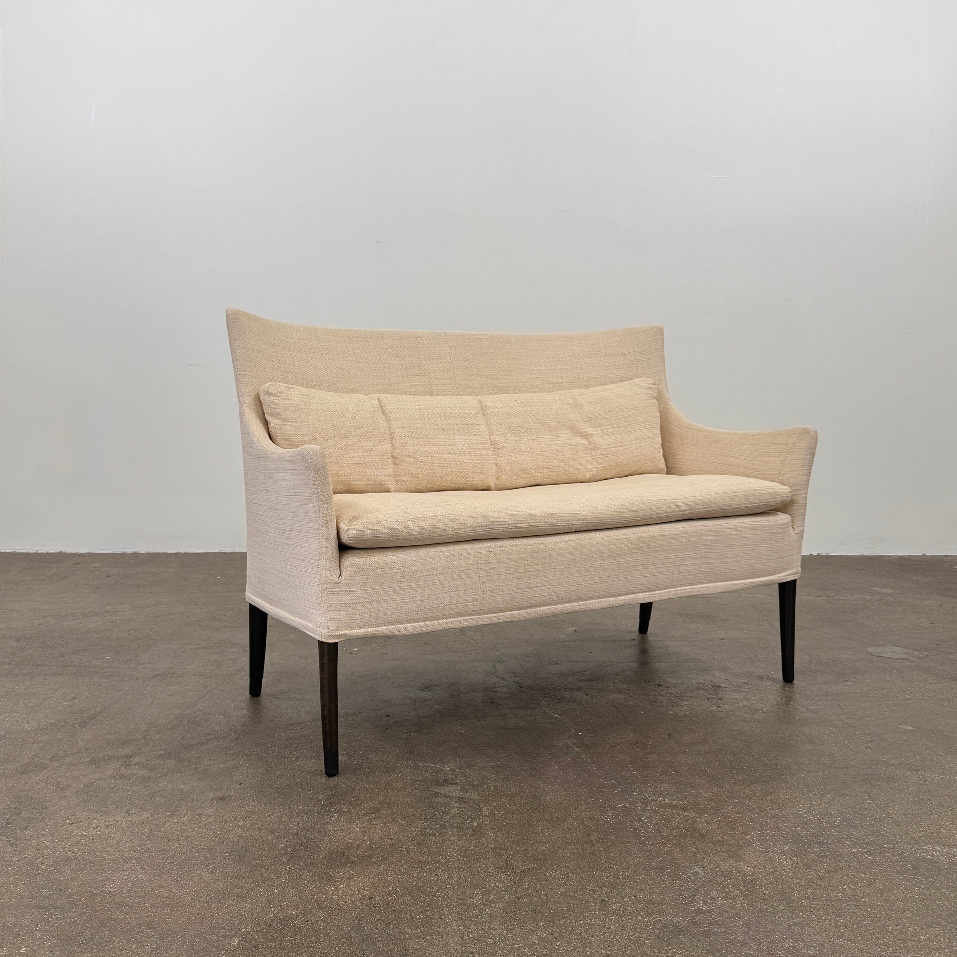 The Giorgetti Settee Sofa by Modern Resale, featuring beige upholstery, two matching cushions, and black legs, is positioned against a plain white wall on a concrete floor.