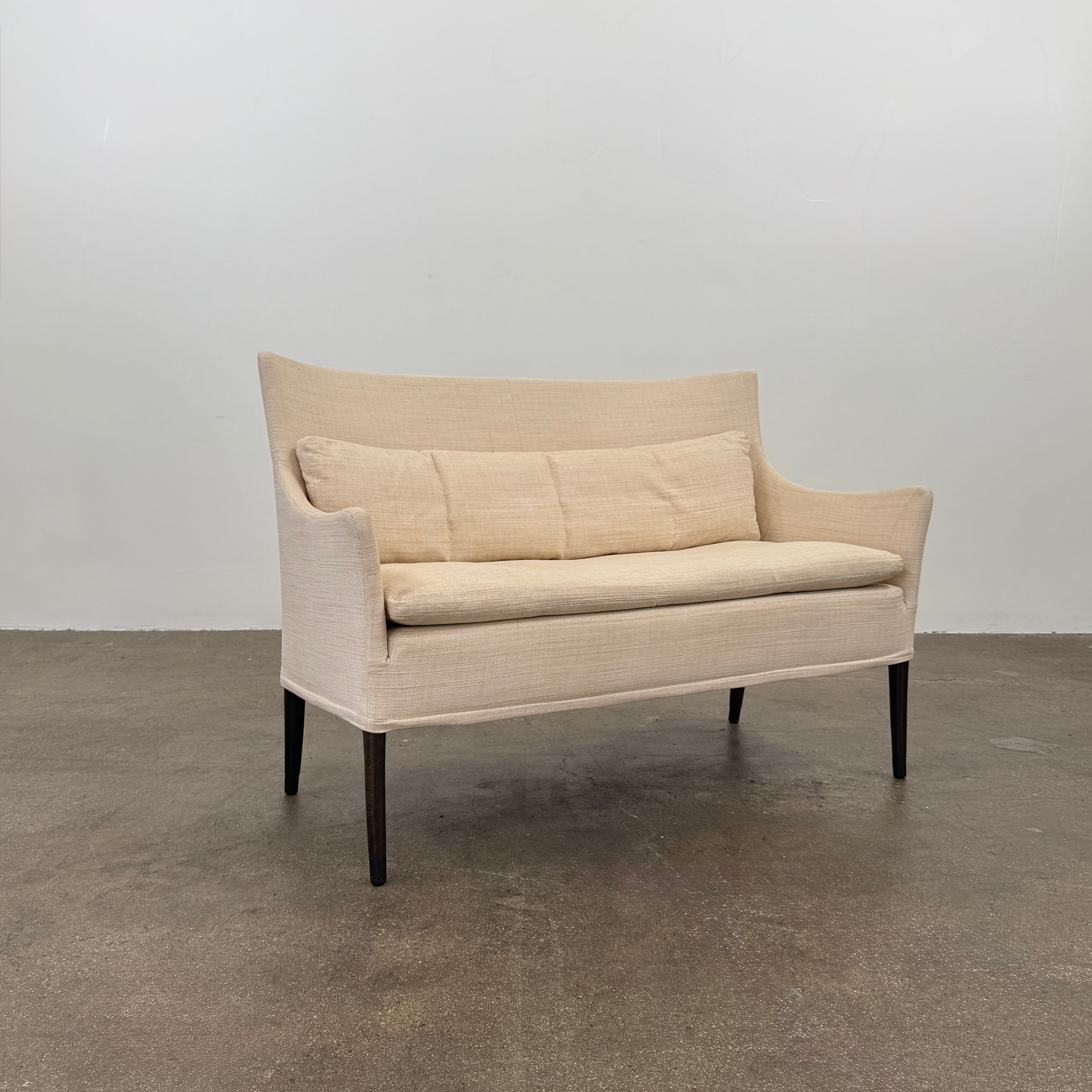 The Giorgetti Settee Sofa by Modern Resale, featuring beige upholstery, two matching cushions, and black legs, is positioned against a plain white wall on a concrete floor.