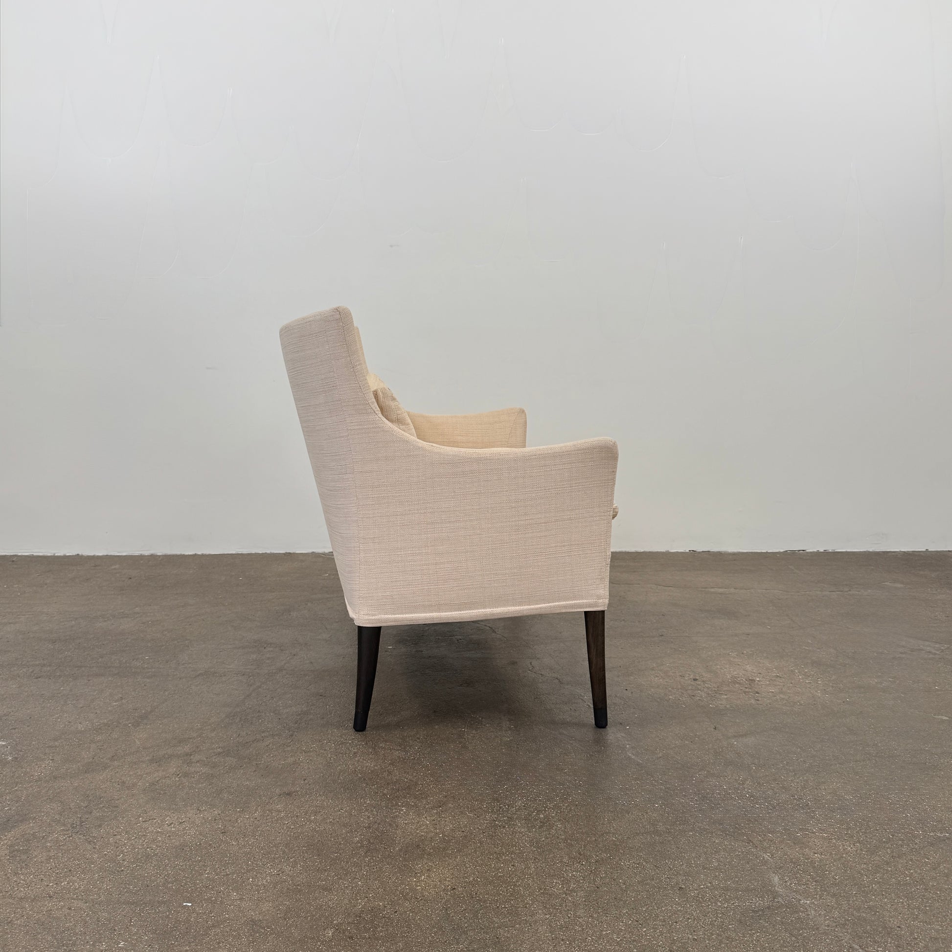 A Vintage Giorgetti Settee by Giorgetti, upholstered in beige with two matching cushions, black legs, and a high back, is placed on a polished concrete floor against a plain white wall.