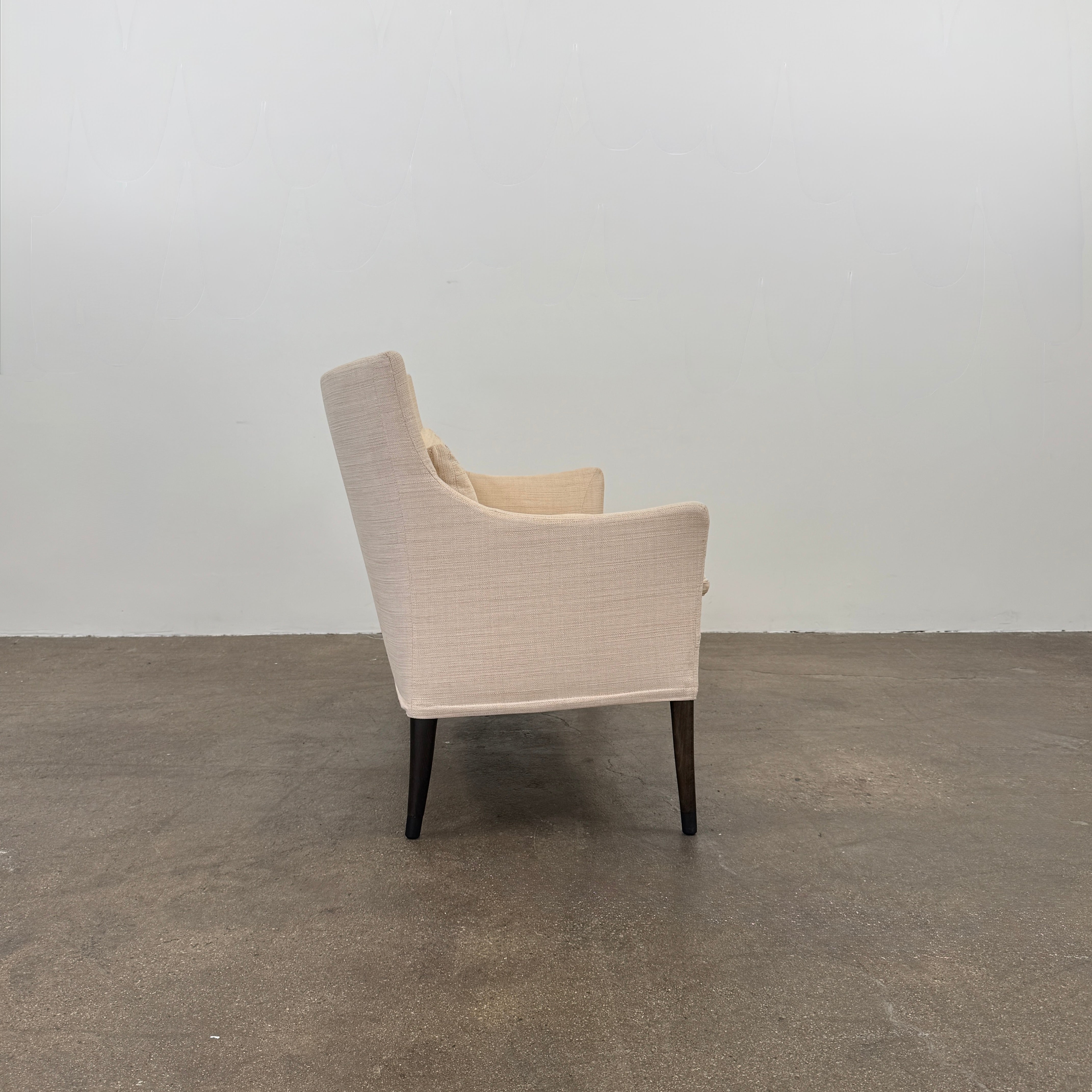 A Vintage Giorgetti Settee by Giorgetti, upholstered in beige with two matching cushions, black legs, and a high back, is placed on a polished concrete floor against a plain white wall.