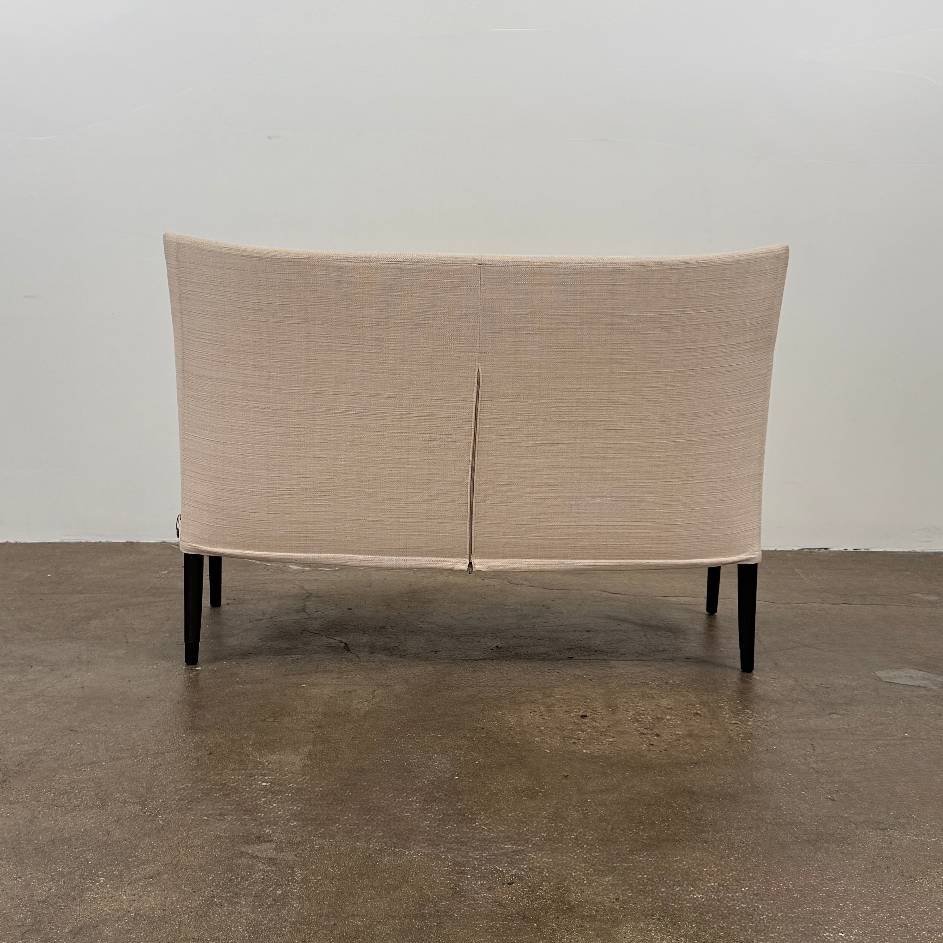 A Vintage Giorgetti Settee by Giorgetti, upholstered in beige with two matching cushions, black legs, and a high back, is placed on a polished concrete floor against a plain white wall.