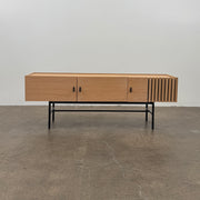 The Woud Array Low Sideboard in white pigmented oak by Modern Resale features three doors, black metal legs, and vertical slats on the right side, displayed against a plain white wall and concrete floor.