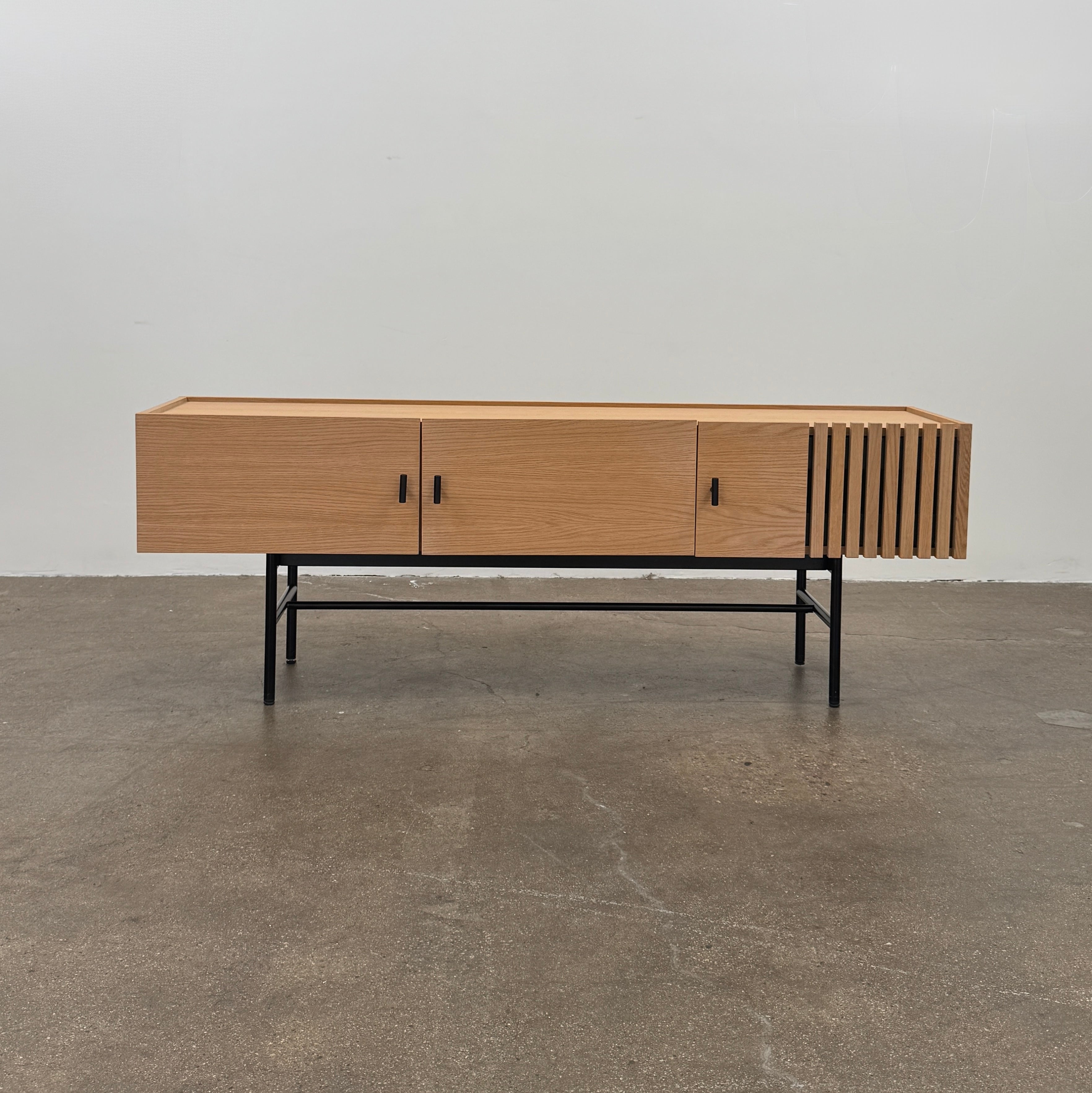 The Woud Array Low Sideboard in white pigmented oak by Modern Resale features three doors, black metal legs, and vertical slats on the right side, displayed against a plain white wall and concrete floor.