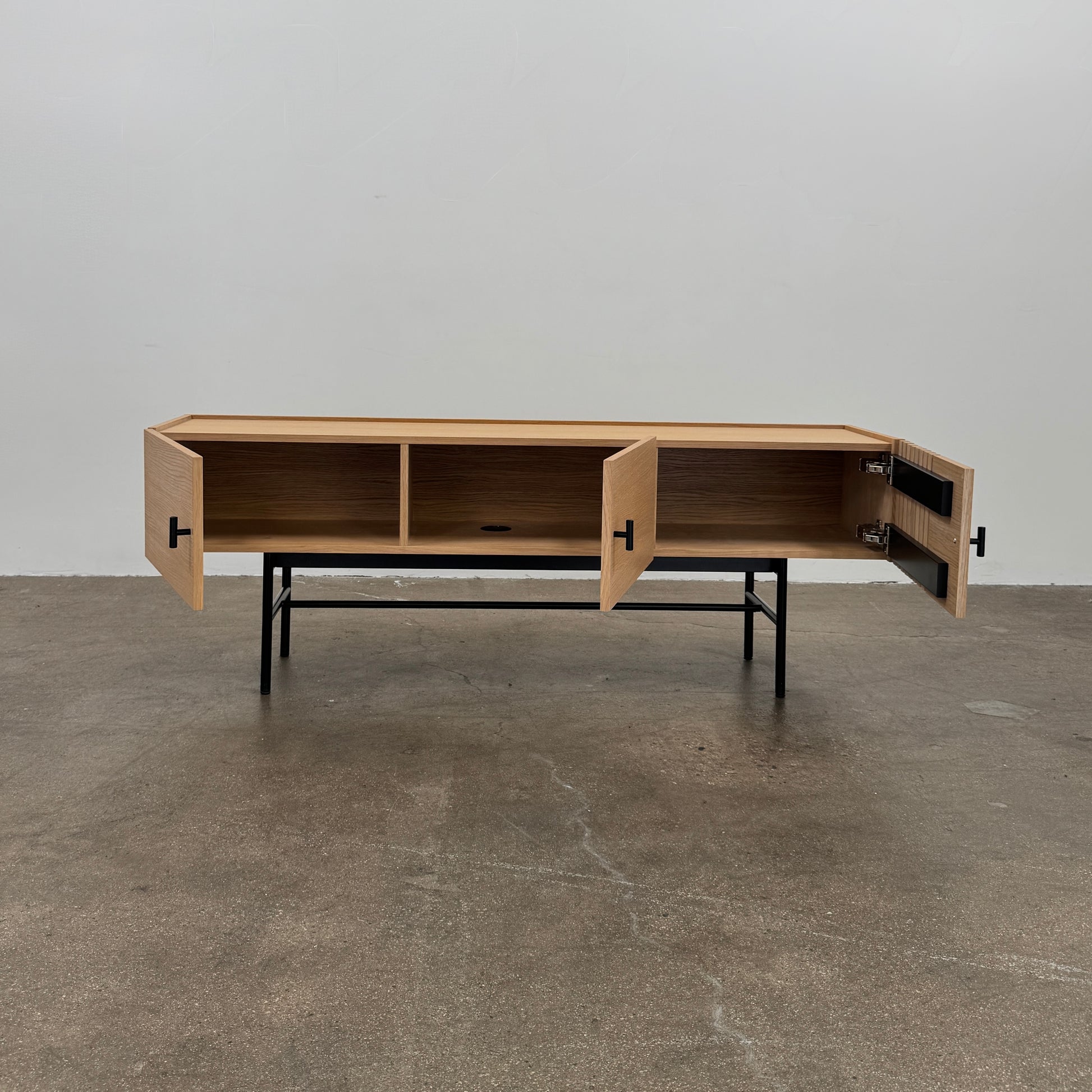 The Woud Array Low Sideboard in white pigmented oak by Modern Resale features three doors, black metal legs, and vertical slats on the right side, displayed against a plain white wall and concrete floor.