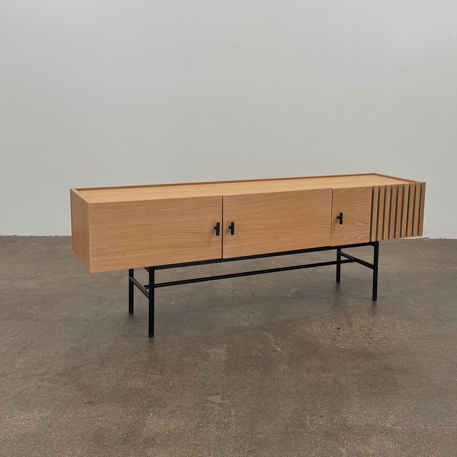 The Woud Array Low Sideboard in white pigmented oak by Modern Resale features three doors, black metal legs, and vertical slats on the right side, displayed against a plain white wall and concrete floor.