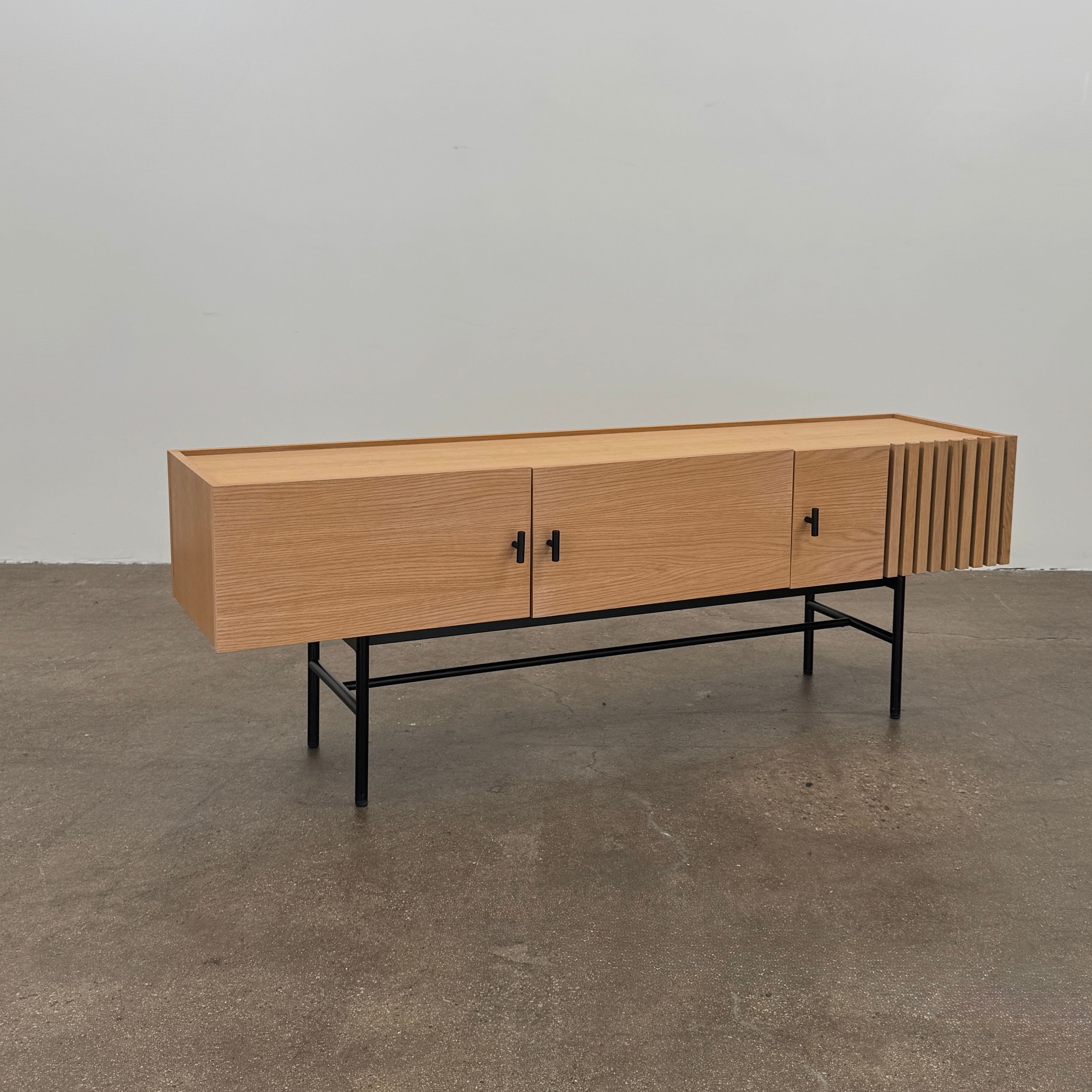 The Woud Array Low Sideboard in white pigmented oak by Modern Resale features three doors, black metal legs, and vertical slats on the right side, displayed against a plain white wall and concrete floor.