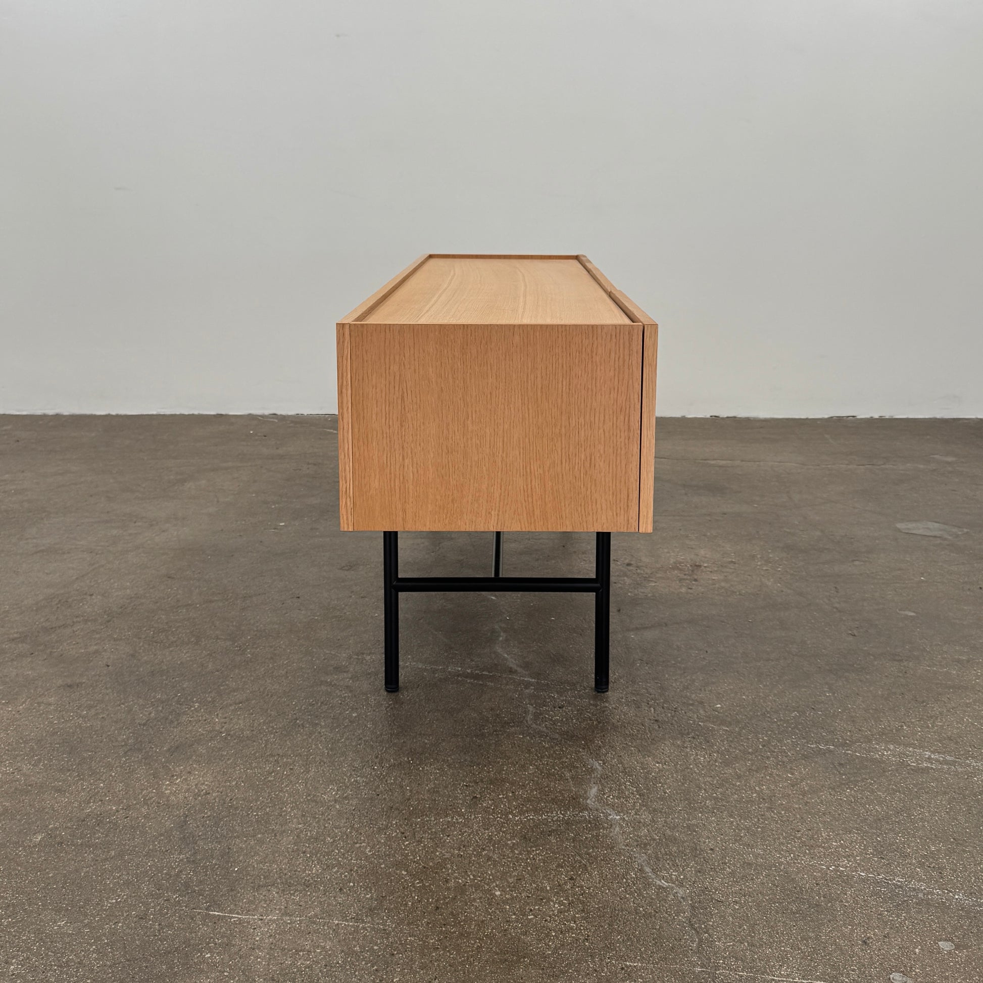 The Woud Array Low Sideboard in white pigmented oak by Modern Resale features three doors, black metal legs, and vertical slats on the right side, displayed against a plain white wall and concrete floor.