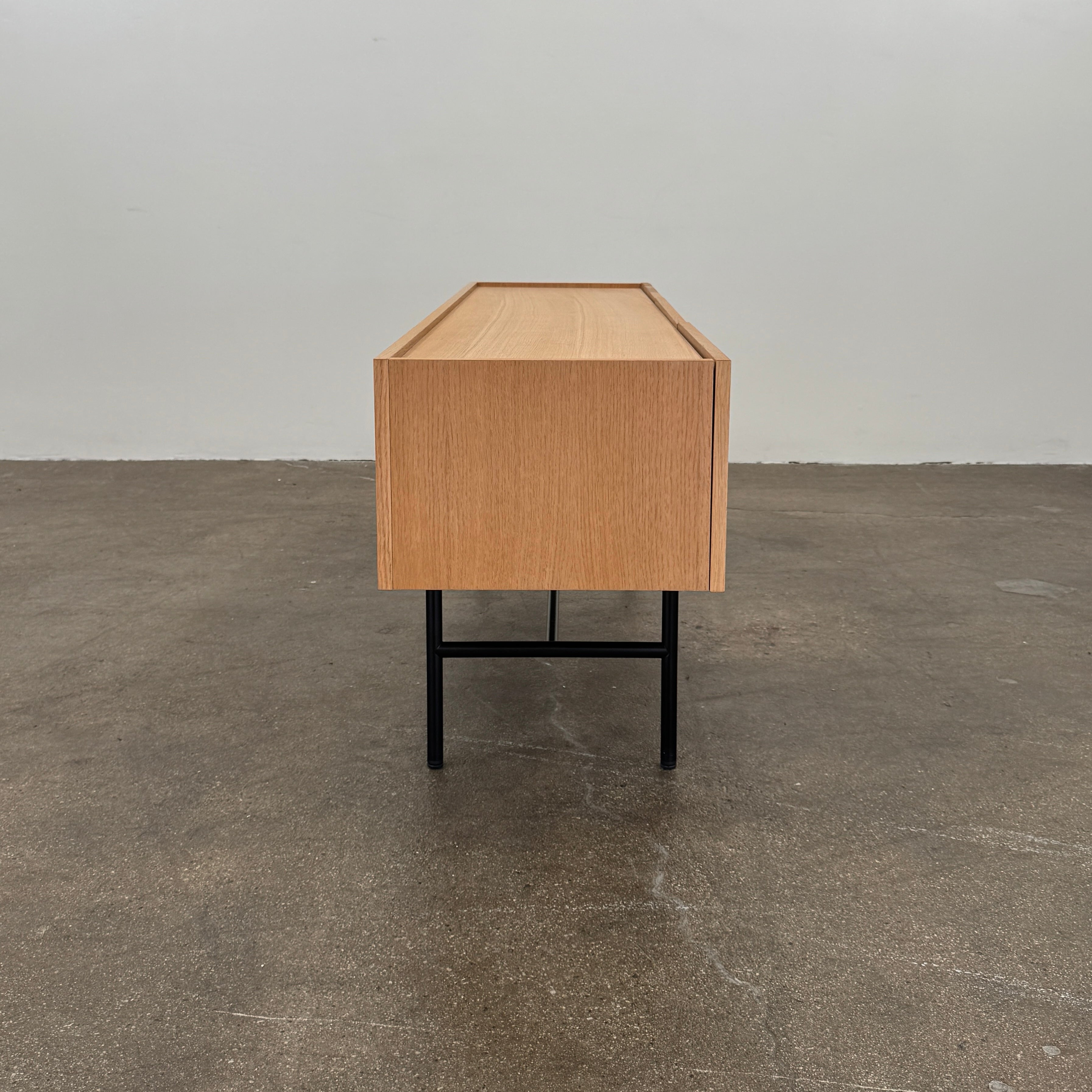 The Woud Array Low Sideboard in white pigmented oak by Modern Resale features three doors, black metal legs, and vertical slats on the right side, displayed against a plain white wall and concrete floor.