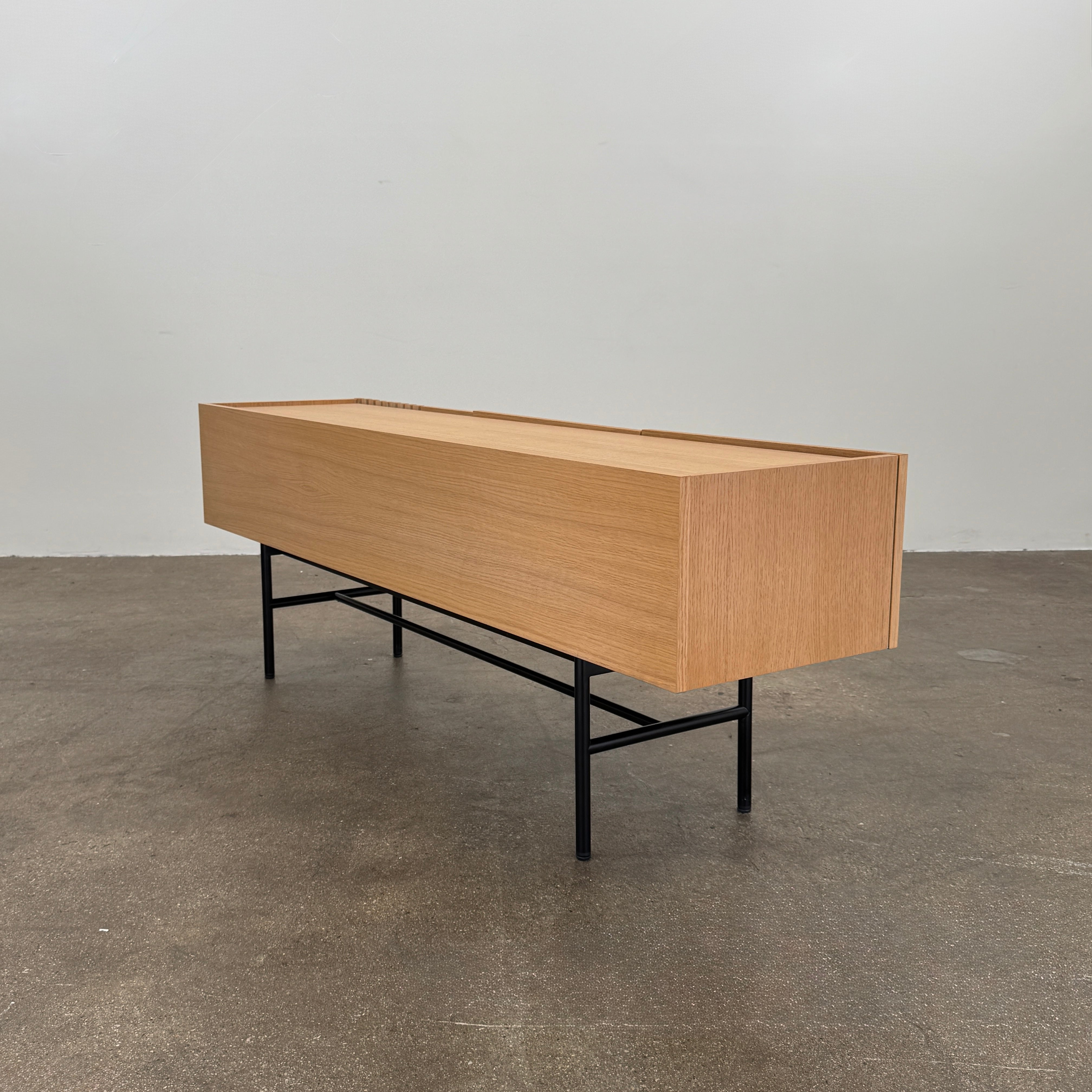 The Woud Array Low Sideboard in white pigmented oak by Modern Resale features three doors, black metal legs, and vertical slats on the right side, displayed against a plain white wall and concrete floor.