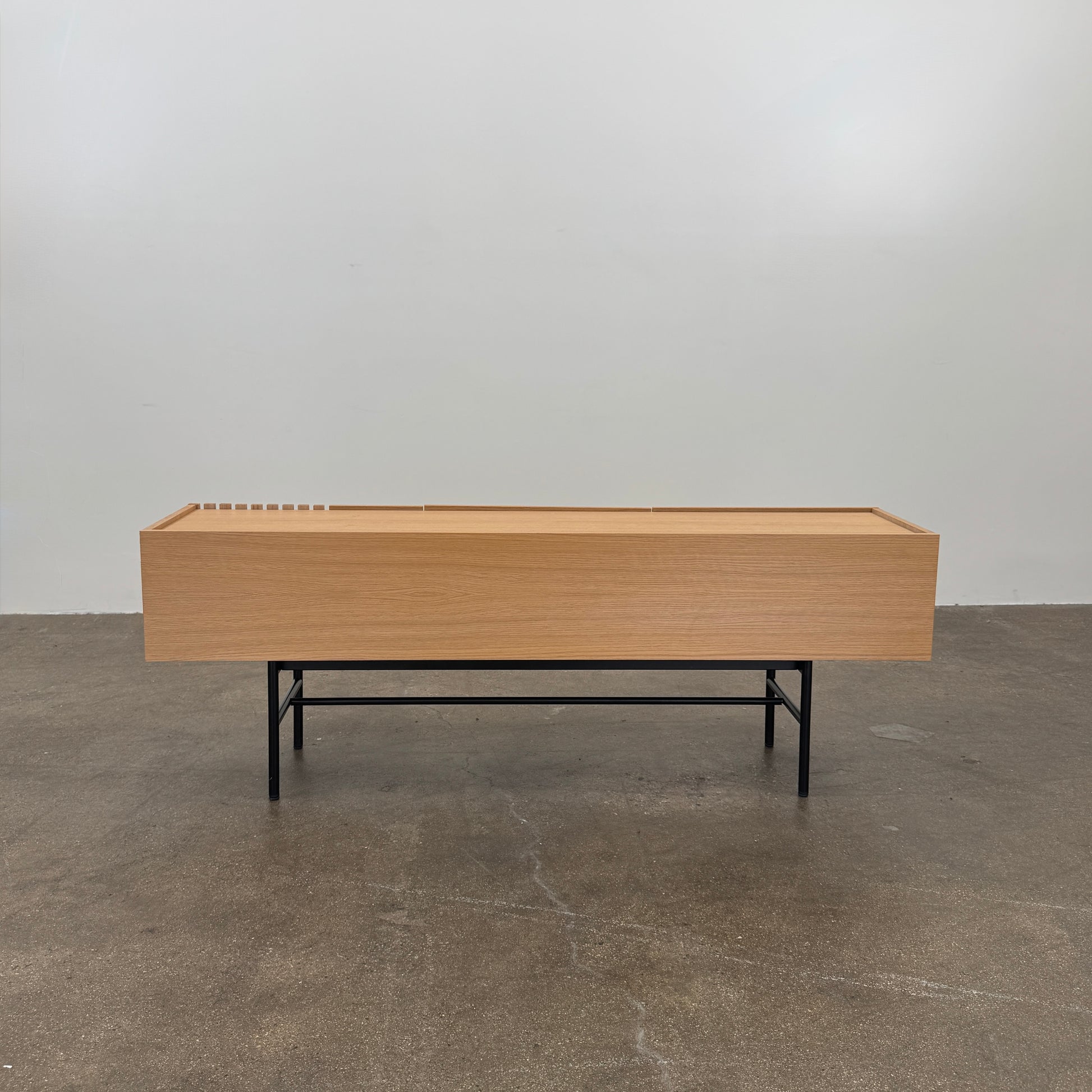 The Woud Array Low Sideboard in white pigmented oak by Modern Resale features three doors, black metal legs, and vertical slats on the right side, displayed against a plain white wall and concrete floor.