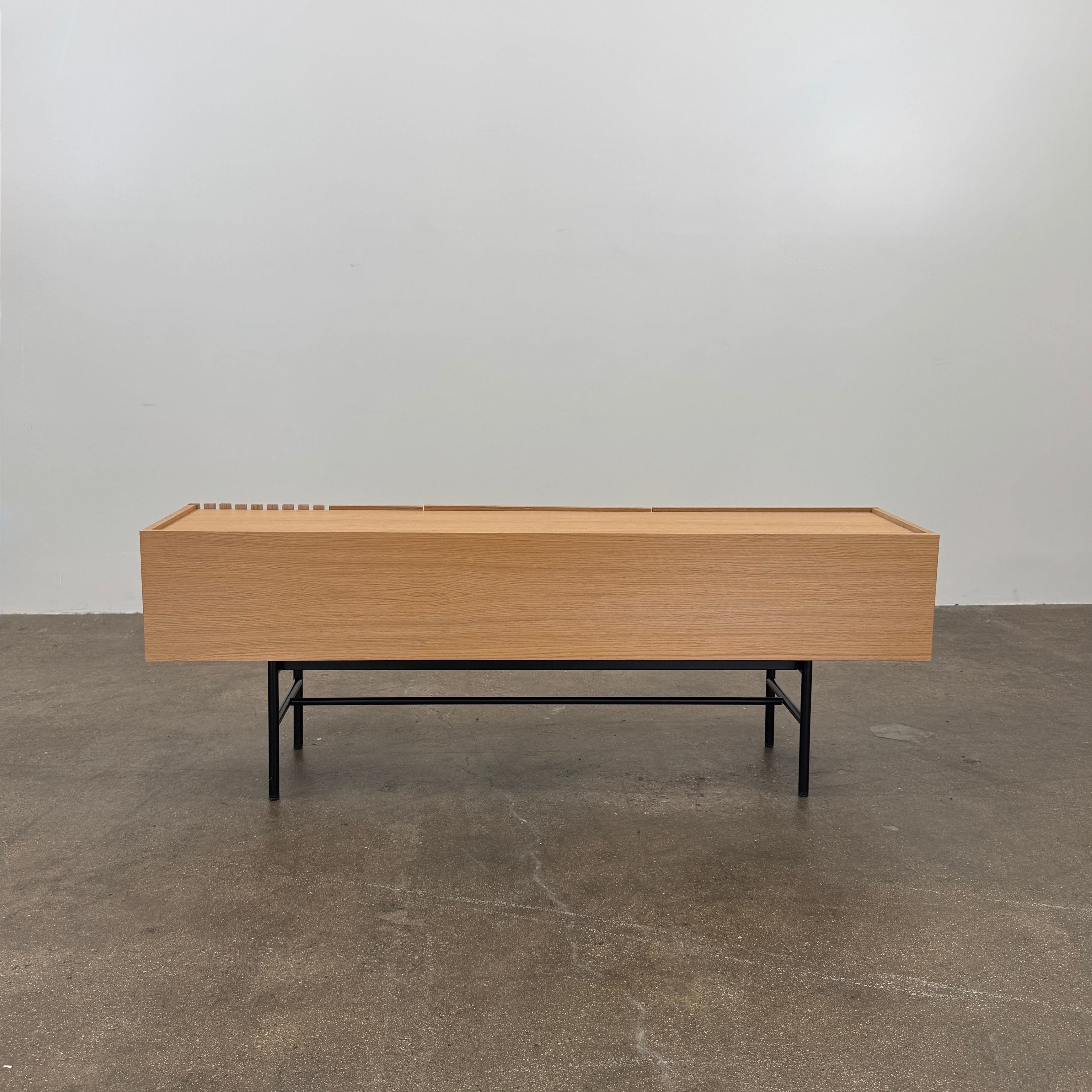 The Woud Array Low Sideboard in white pigmented oak by Modern Resale features three doors, black metal legs, and vertical slats on the right side, displayed against a plain white wall and concrete floor.