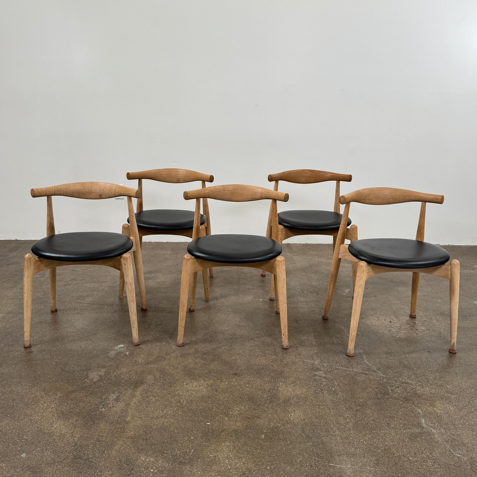 A set of five Carl Hansen & Son CH20 Elbow Chairs by Carl Hansen & Son, each with a curved wooden backrest and a round black seat cushion, are arranged in two rows against a plain white background.